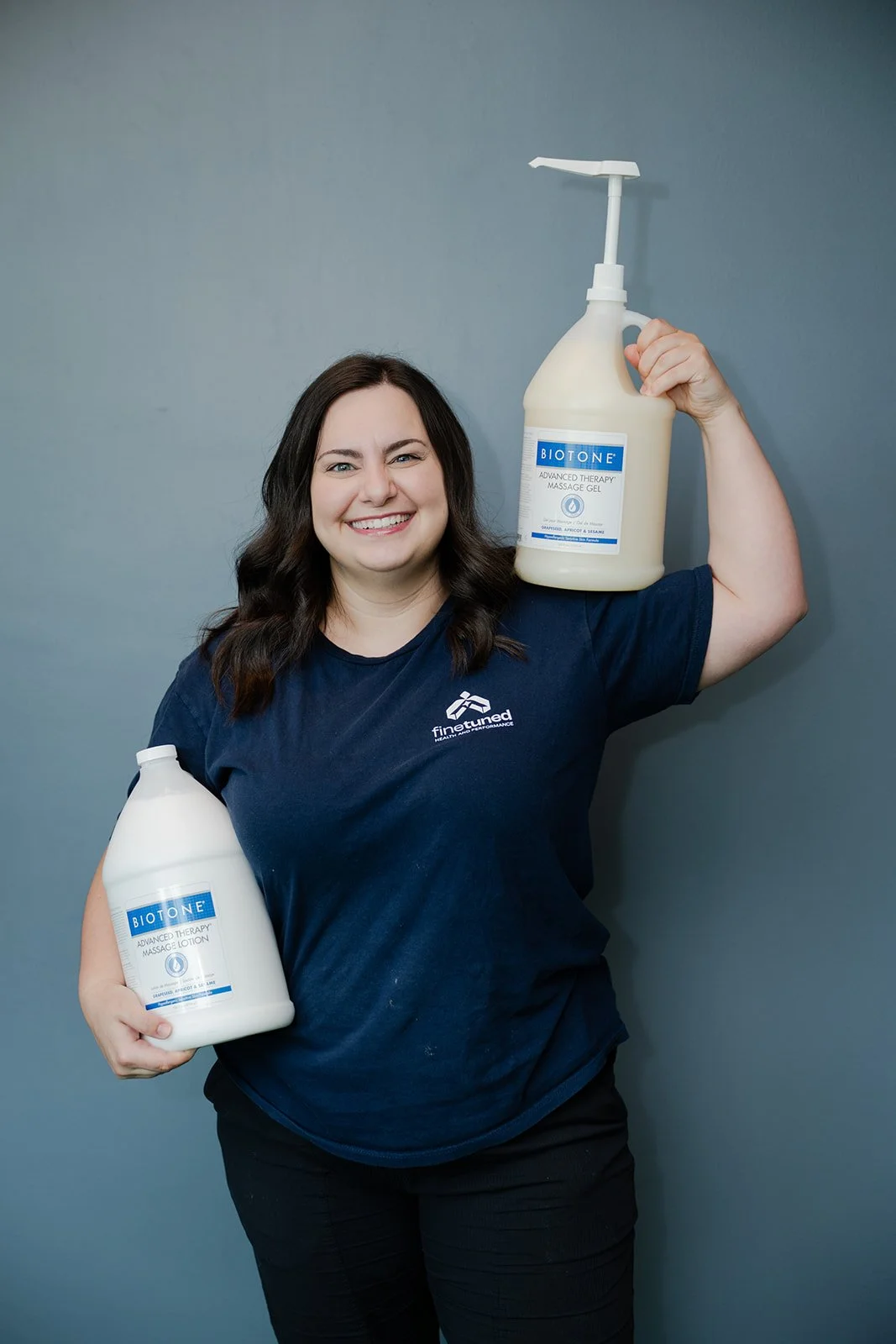 A woman with long dark hair smiling, holding two large bottles of Biotone massage gel, wearing a navy blue T-shirt with a logo on it, standing against a plain grey wall.