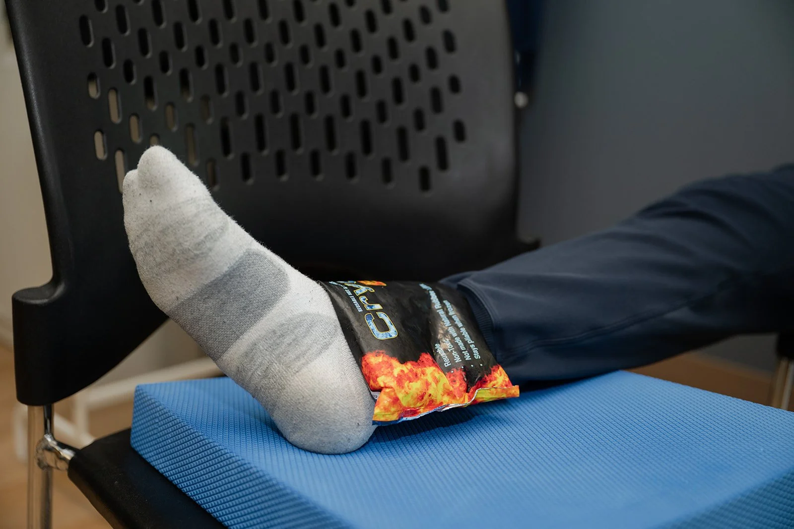 A person sitting on a wooden bench, putting on a white sock with red stripes near the top, wearing white sneakers, with their legs and hands visible, and wearing a beige and orange striped sock on one foot.