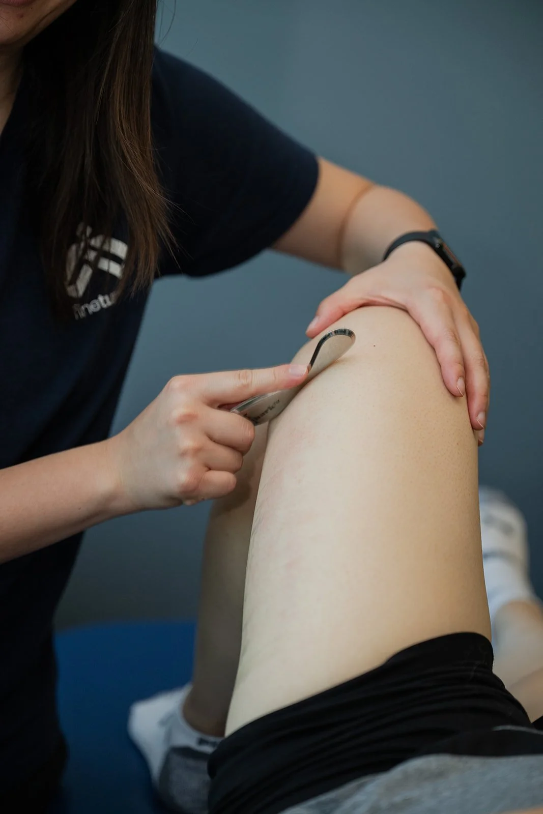 A female chiropractowearing a black long sleeve shirt and gray pants is performing a hip joint mobilisation of a patient's leg, who is wearing black shorts. They are in a room with anatomical posters of the skeletal and muscular systems on the wall.