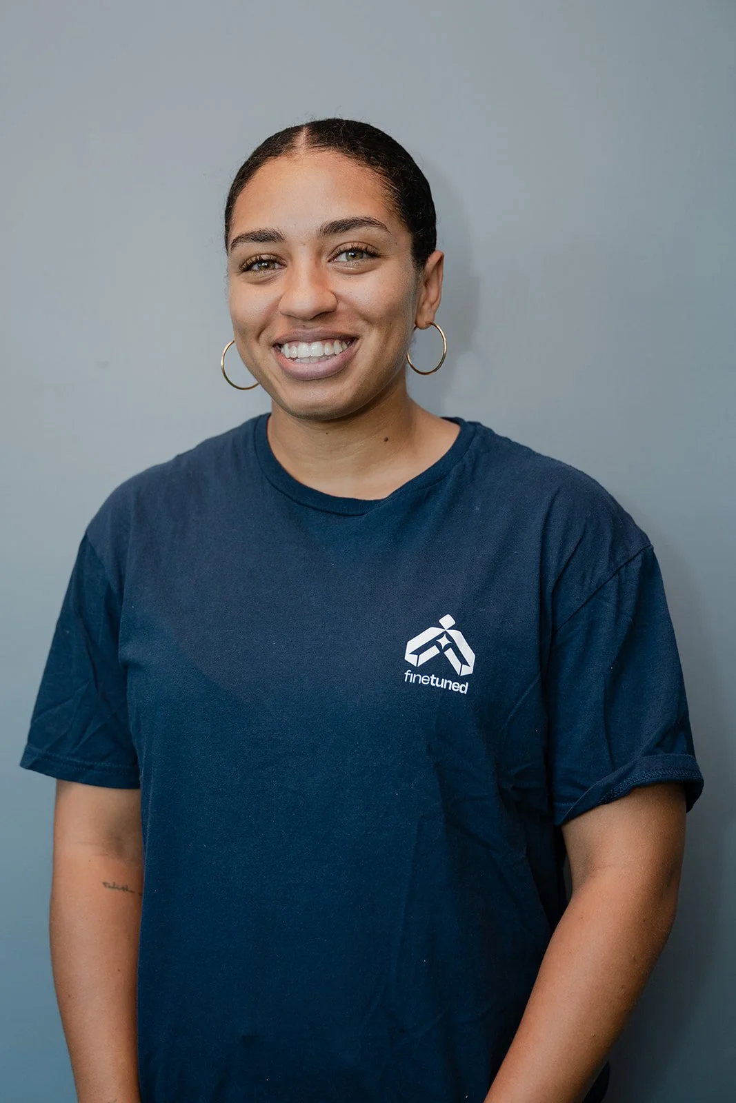 A smiling woman with braided hair wearing a navy blue t-shirt with a Finetuned logo, standing against a plain gray background.