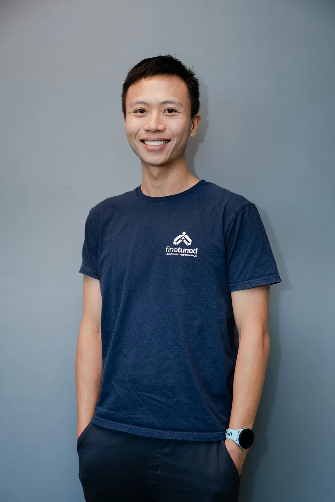 A person wearing a navy blue t-shirt with a logo and the words "finetuned health and performance" standing in front of a light blue wall.