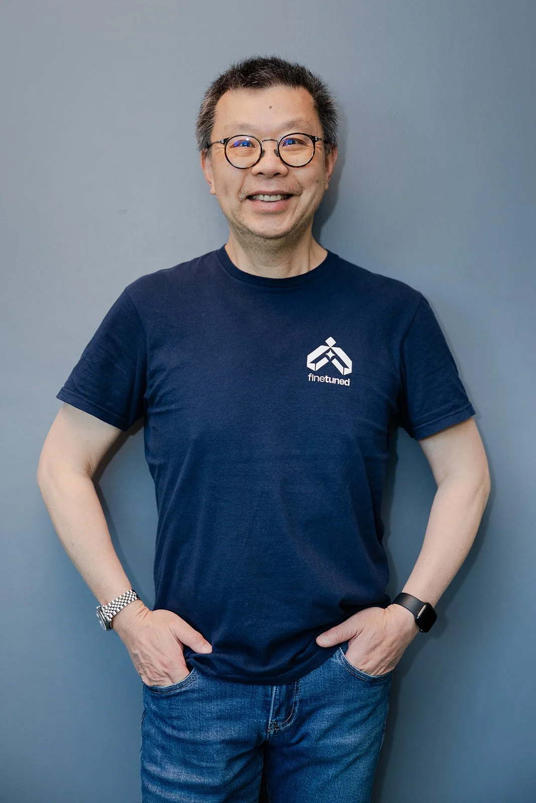 A smiling man with glasses, wearing a navy blue t-shirt with a 'finetuned' logo, standing with hands in his pockets against a gray wall.