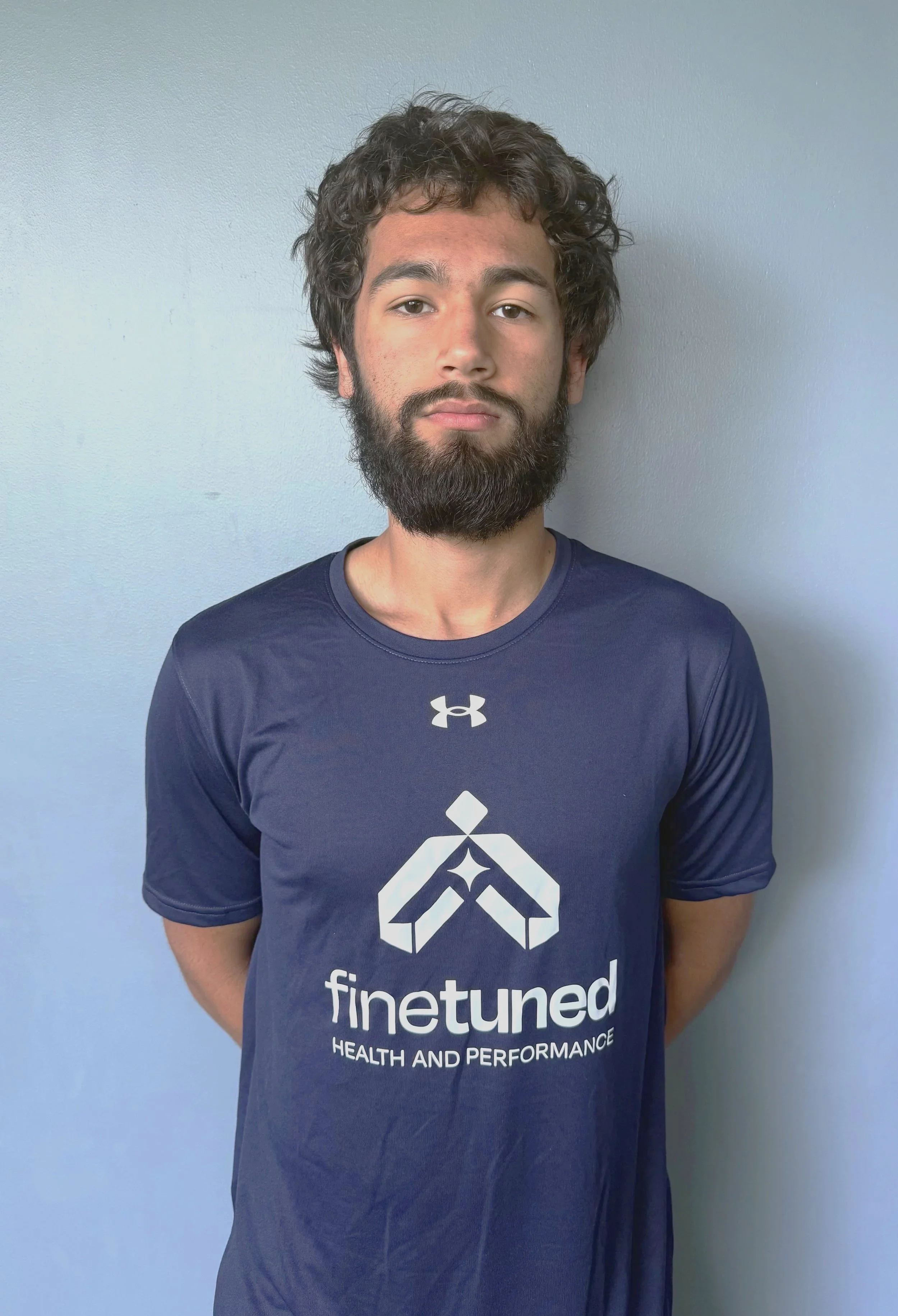 A young man with curly hair and a beard stands against a light blue wall, wearing a navy blue Finetuned shirt with the company's logo and slogan.