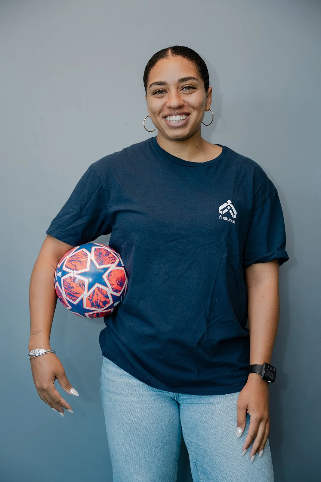 A woman with dark hair, wearing hoop earrings, a navy blue t-shirt, light jeans, and a fitness tracker, holding a colorful soccer ball with a star pattern, standing against a plain gray wall.