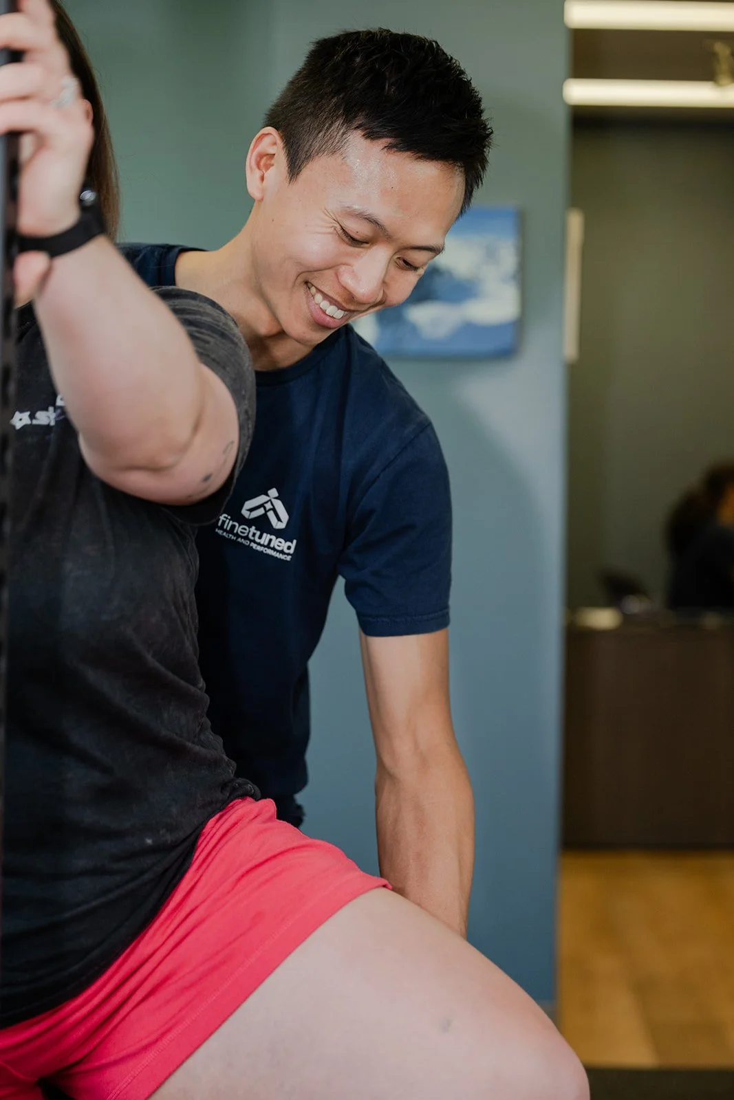 A smiling man in a blue t-shirt assisting another person in a black shirt with a red pair of shorts during a physical therapy or fitness session indoors.