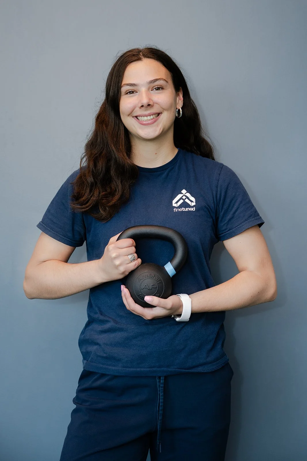 A woman with long dark hair smiling and holding a kettlebell against a plain gray background. She is wearing a navy blue t-shirt with a logo and text that says 'fined tuned,' and navy blue pants.