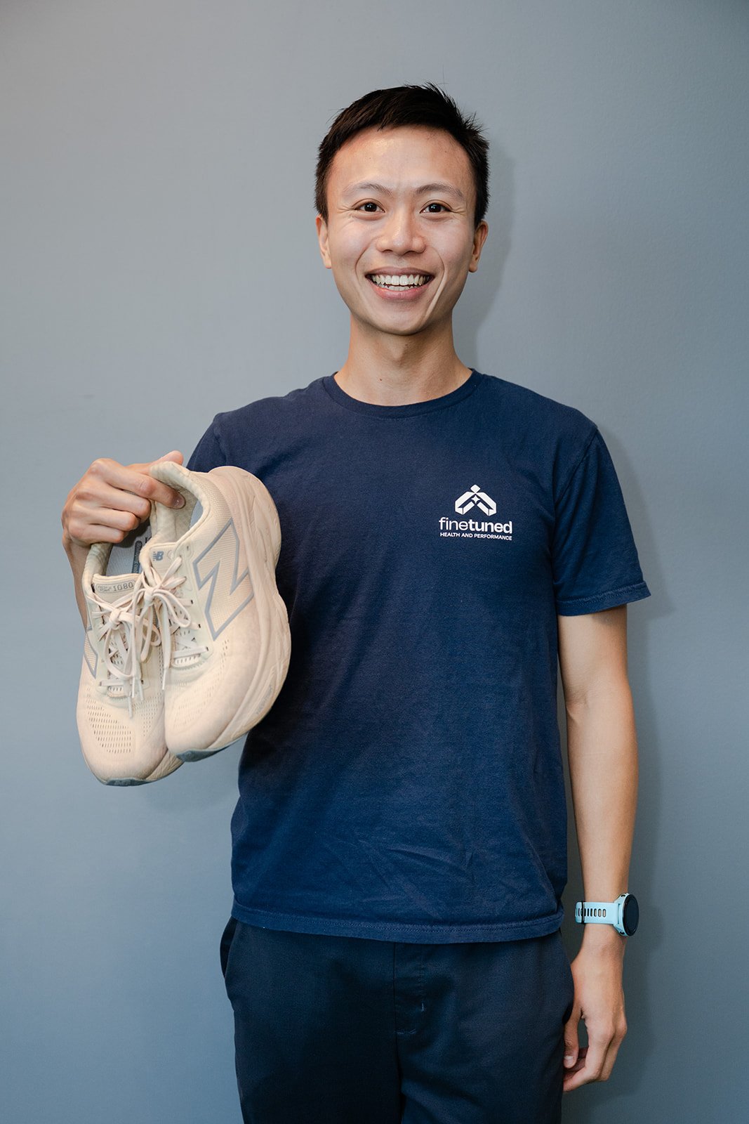 A man smiling and holding a beige running shoe in his right hand. He wears a dark blue t-shirt with a logo and the text 'finetuned' and 'Health and Performance', and a white wristwatch, standing against a gray background.
