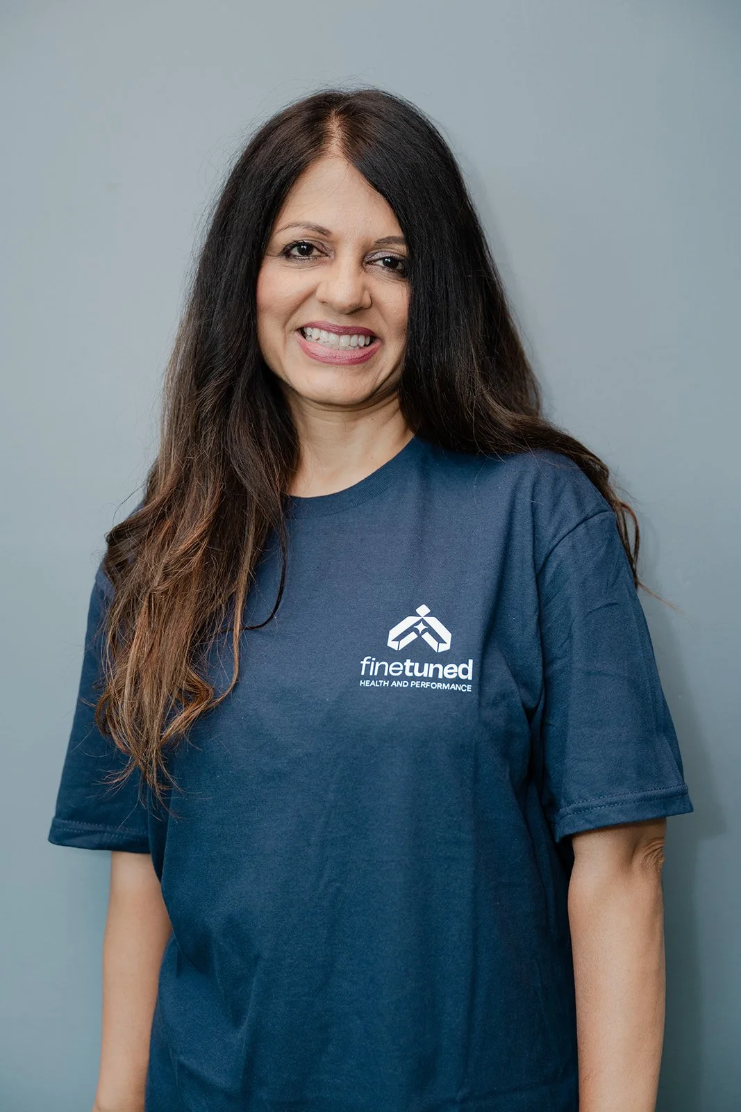 Portrait of a woman with long dark hair, smiling, wearing a black zip-up jacket with a logo on the chest, standing against a plain light blue wall.