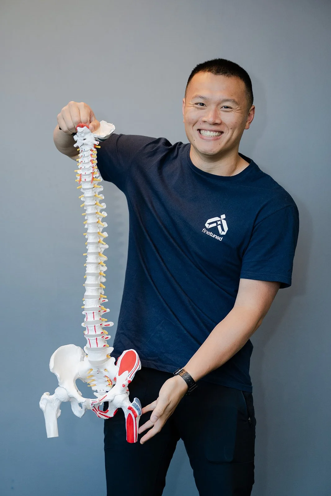 A person smiling and holding a model of a human spine with pelvis against a gray background.