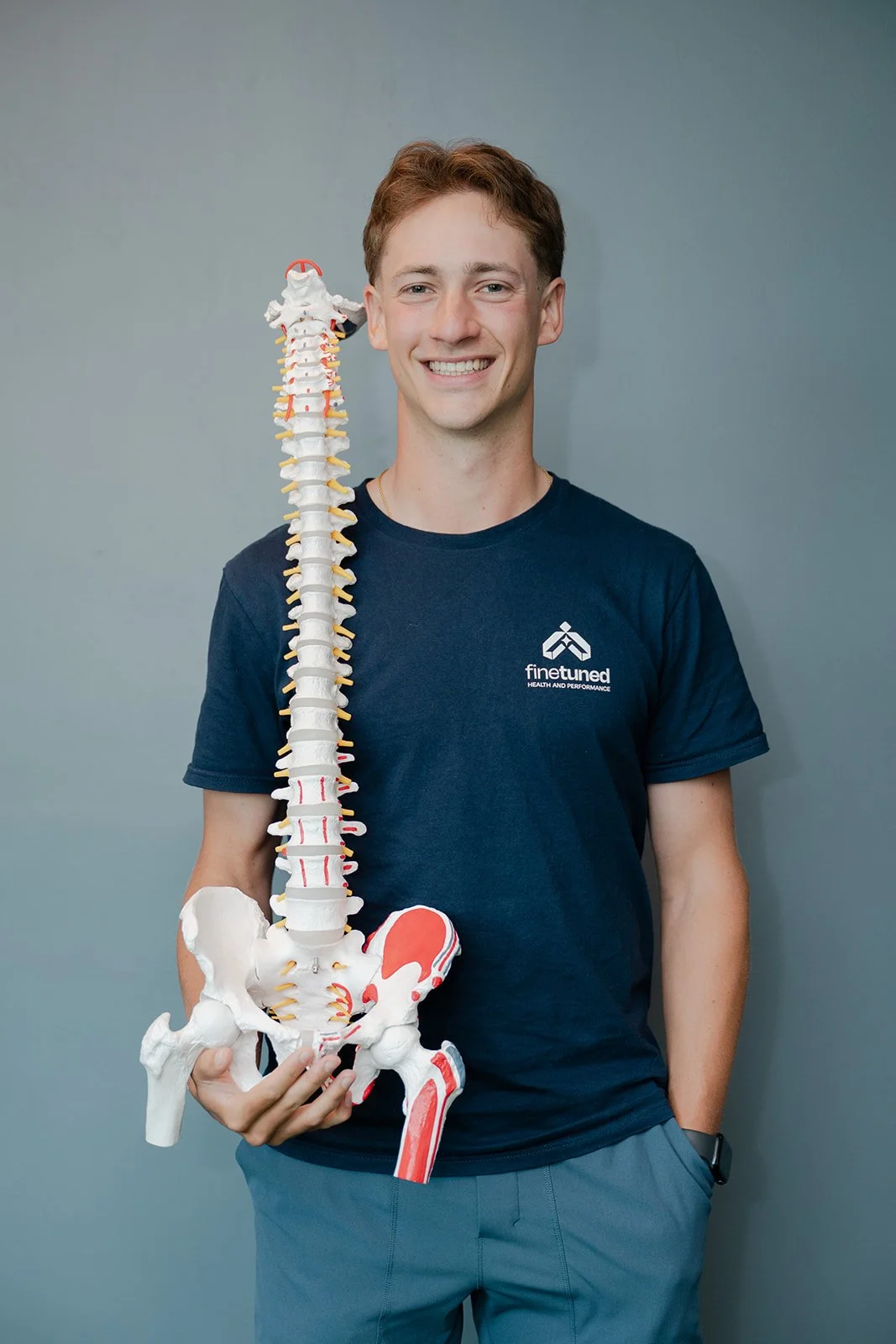 Young man smiling holding a medical model of the human spine and pelvis, standing against a plain wall.