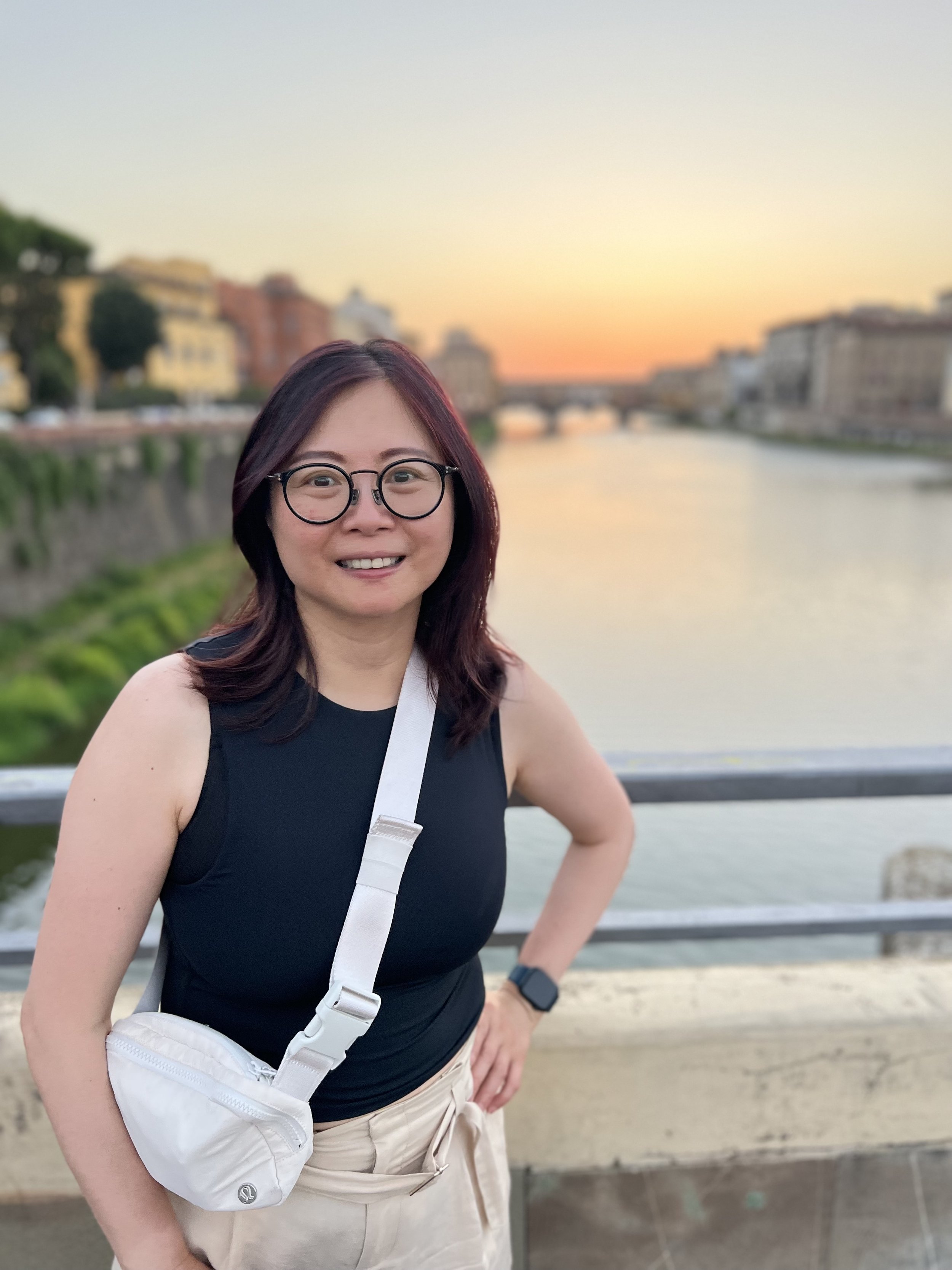 A woman with shoulder-length dark red hair and glasses smiling at the camera, standing outdoors near a river with a cityscape and a bridge in the background during sunset.