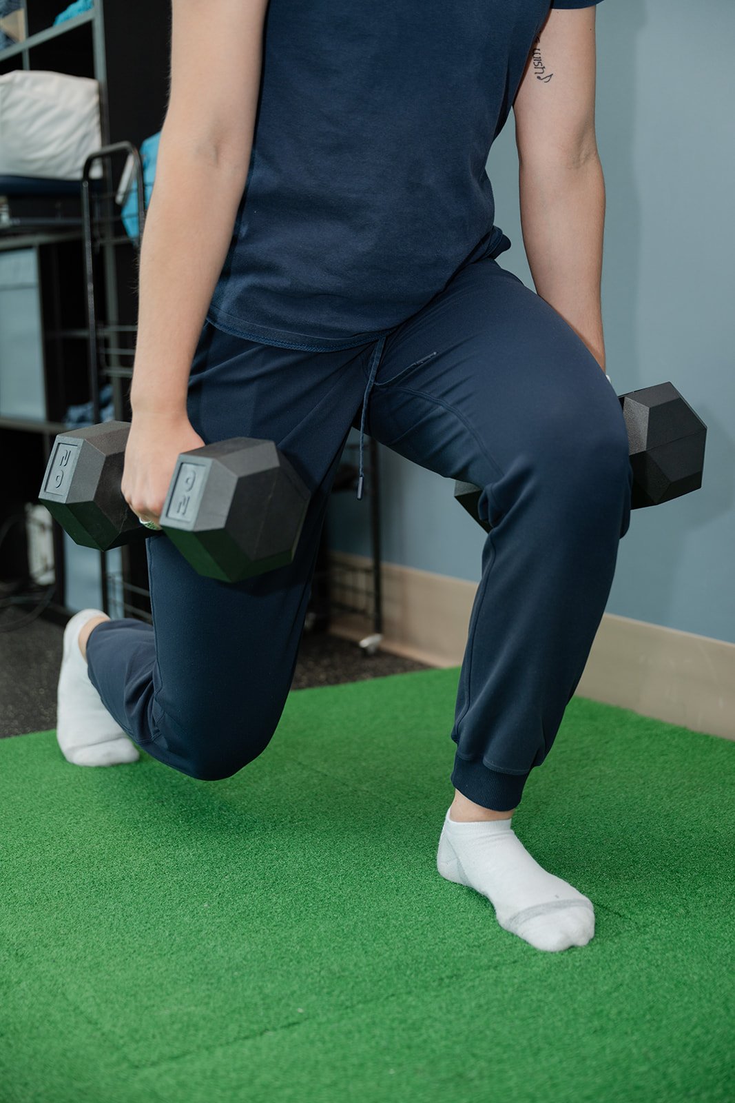 Two people lifting blue kettlebells in a gym with a red wall and wooden panels in the background.