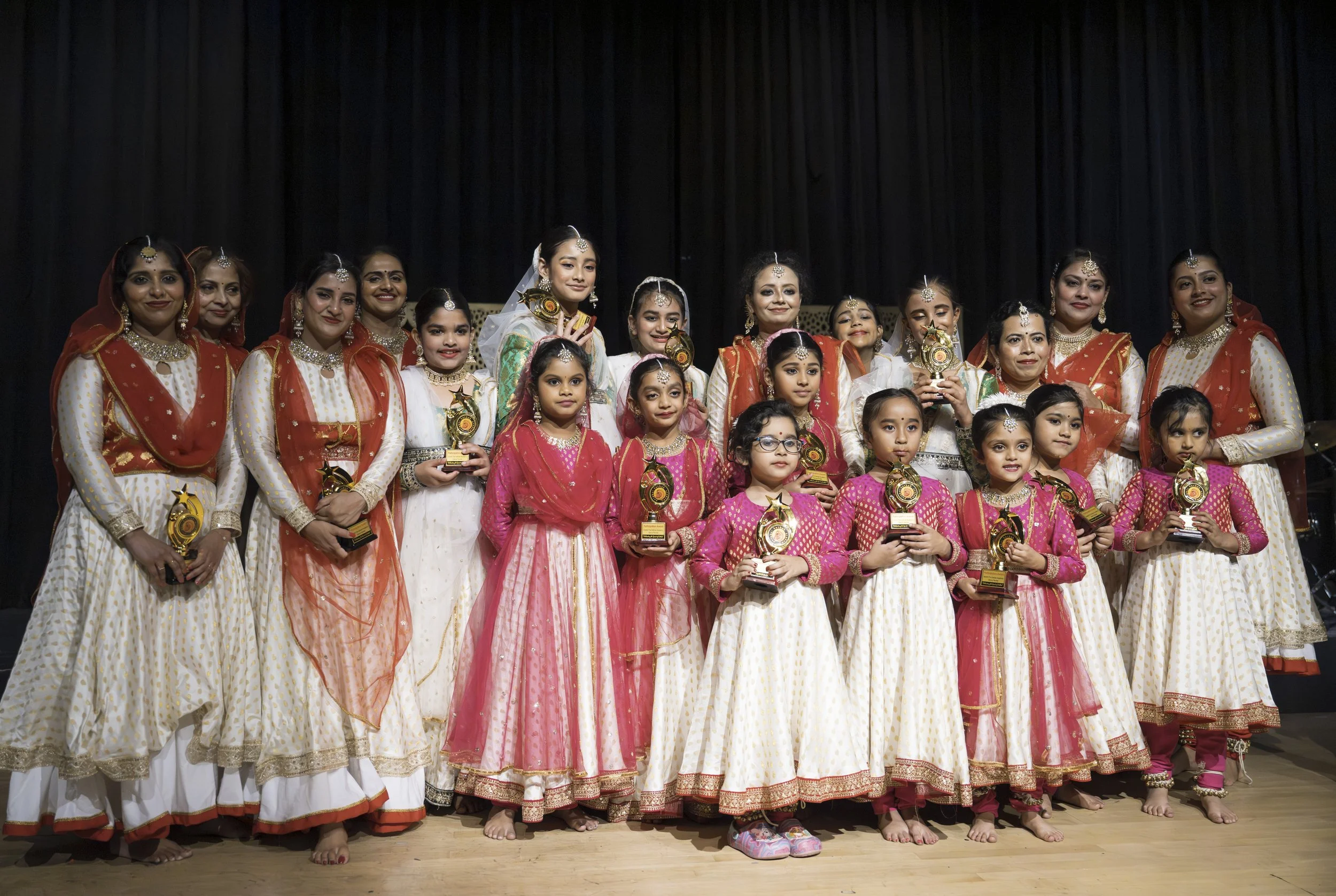 A group of women and girls dressed in traditional Indian attire, holding trophies, standing on a stage with a black curtain backdrop.