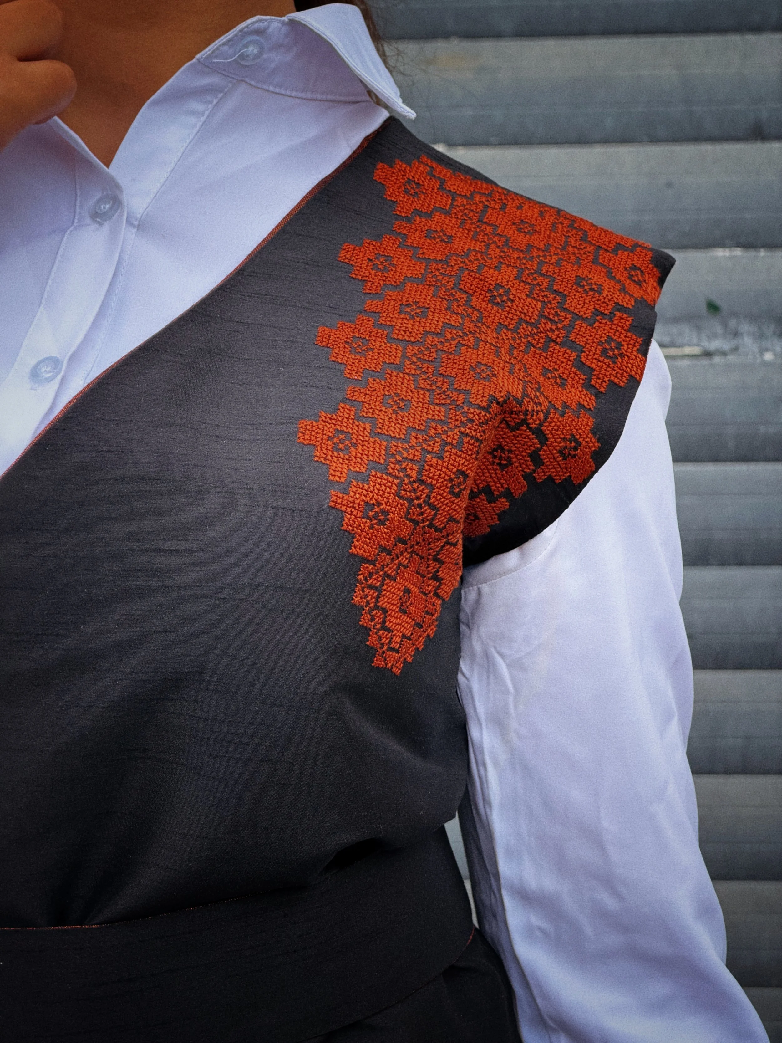 Close-up of a black taffeta asymmetric waistcoat featuring orange Palestinian tatreez embroidery across the shoulder, worn over a white shirt.