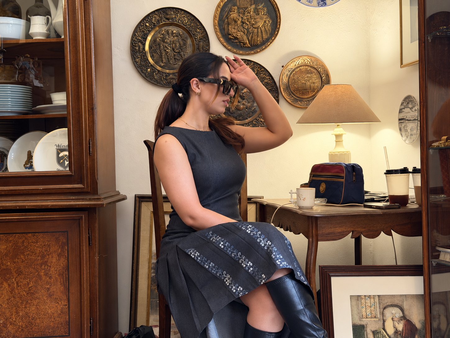 Model seated in a charcoal sleeveless denim dress featuring a fitted bodice, pleated skirt and traditional Palestinian tatreez hand embroidery.