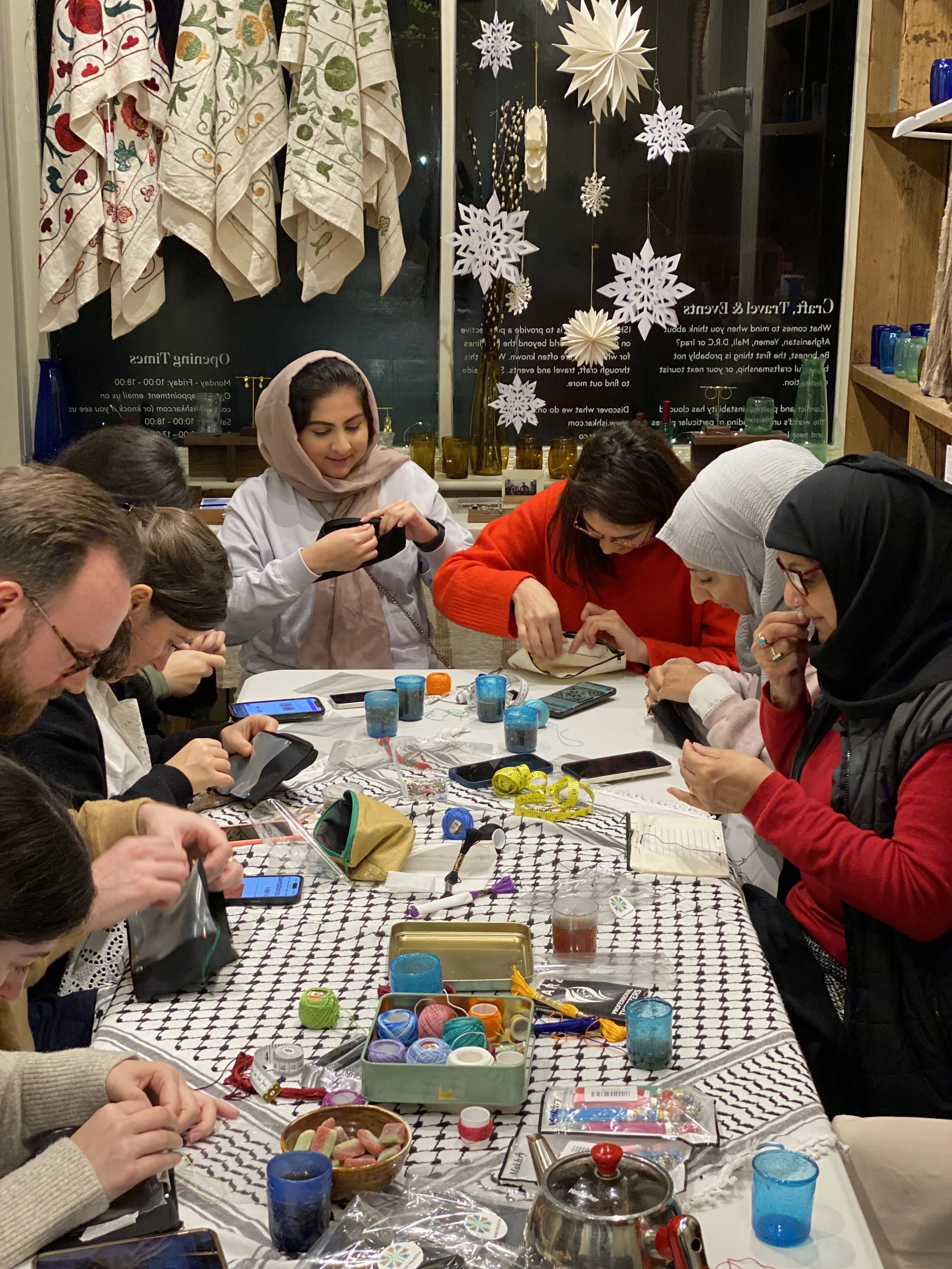 Group of people sitting around a table engaged in a craft activity, with various craft supplies and cell phones on the table, in a room decorated with holiday ornaments and snowflake decorations.
