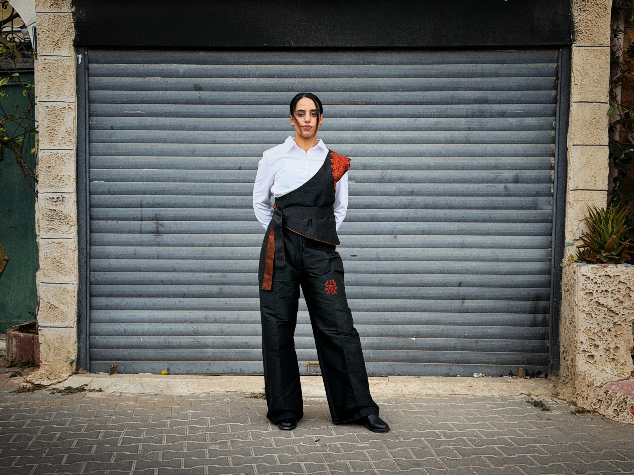 Full-length model wearing a black Palestinian tatreez embroidered waistcoat with matching wide-leg trousers and a white shirt.