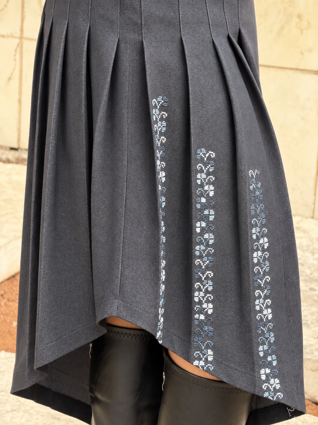 Close-up of blue and white Palestinian tatreez embroidery along the pleats of a charcoal cotton denim dress with an asymmetric hem.