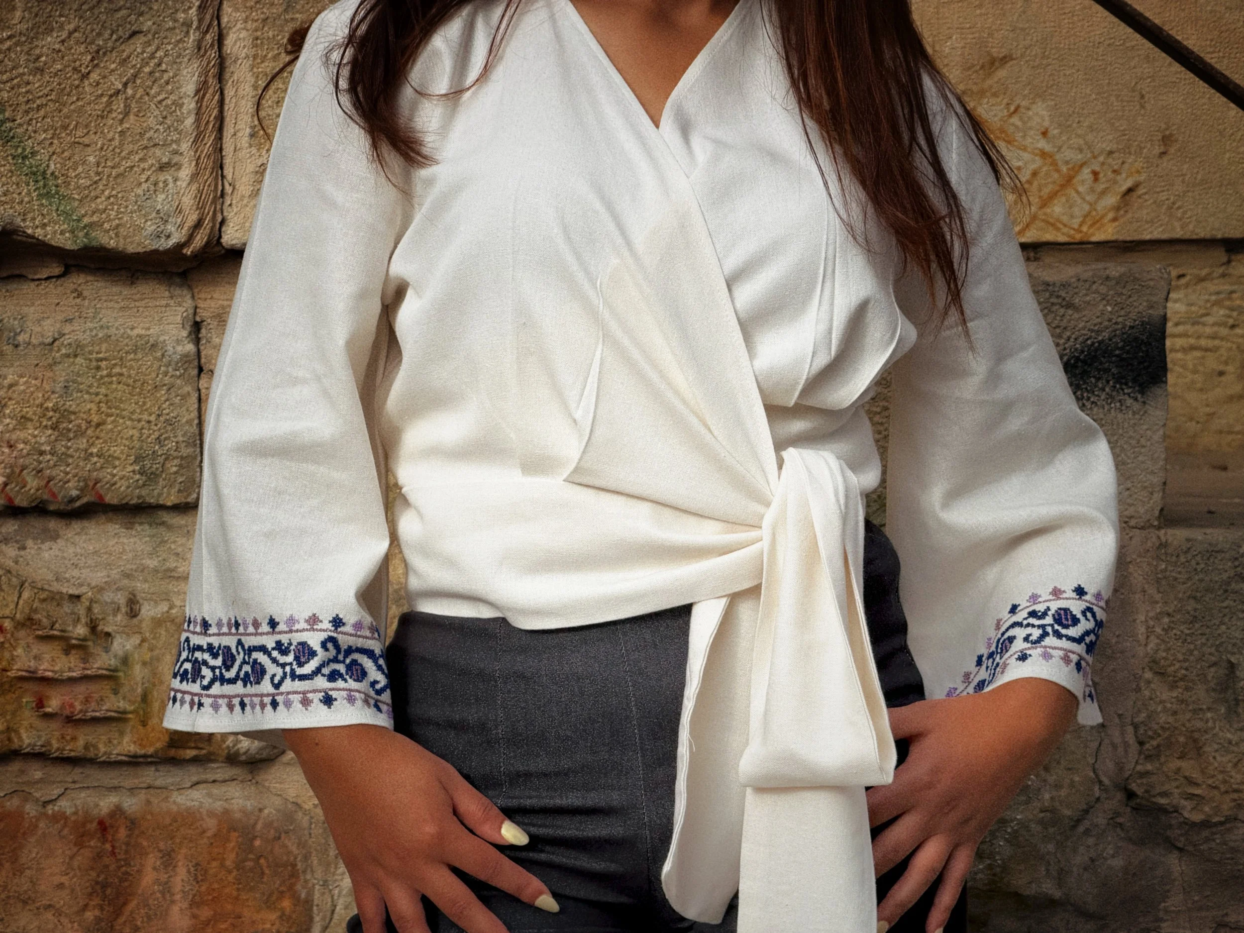 Model wearing an ivory wrap blouse with blue and purple Palestinian tatreez embroidered cuffs and a side tie, standing beside a dark doorway.