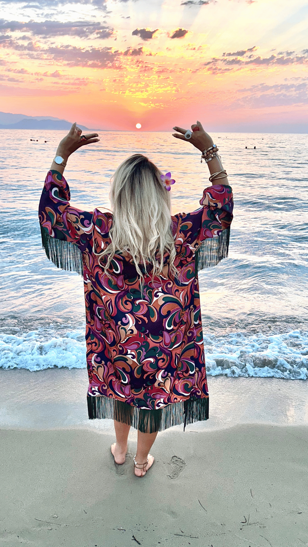 Personne de dos sur une plage au coucher du soleil, portant une robe à motifs, les bras levés.