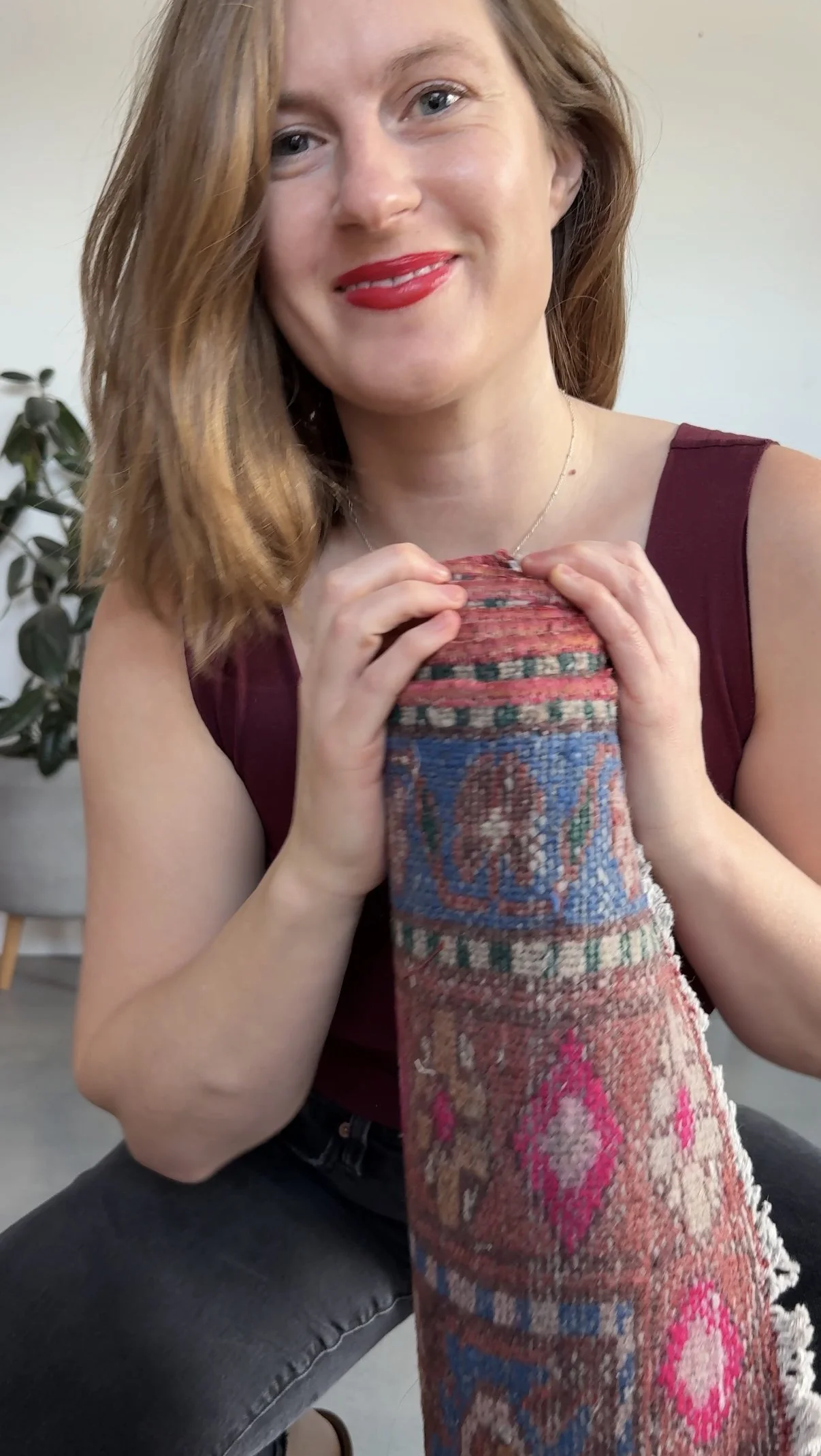 A smiling woman with red hair, wearing red lipstick and a sleeveless maroon top, holding a colorful patterned fabric or textile in front of her chest.