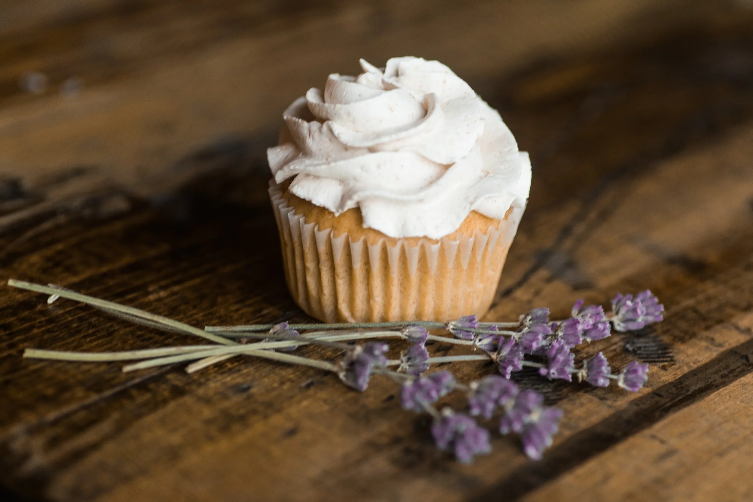 Cupcake with white frosting next to sprigs of purple flowers on a wooden table.