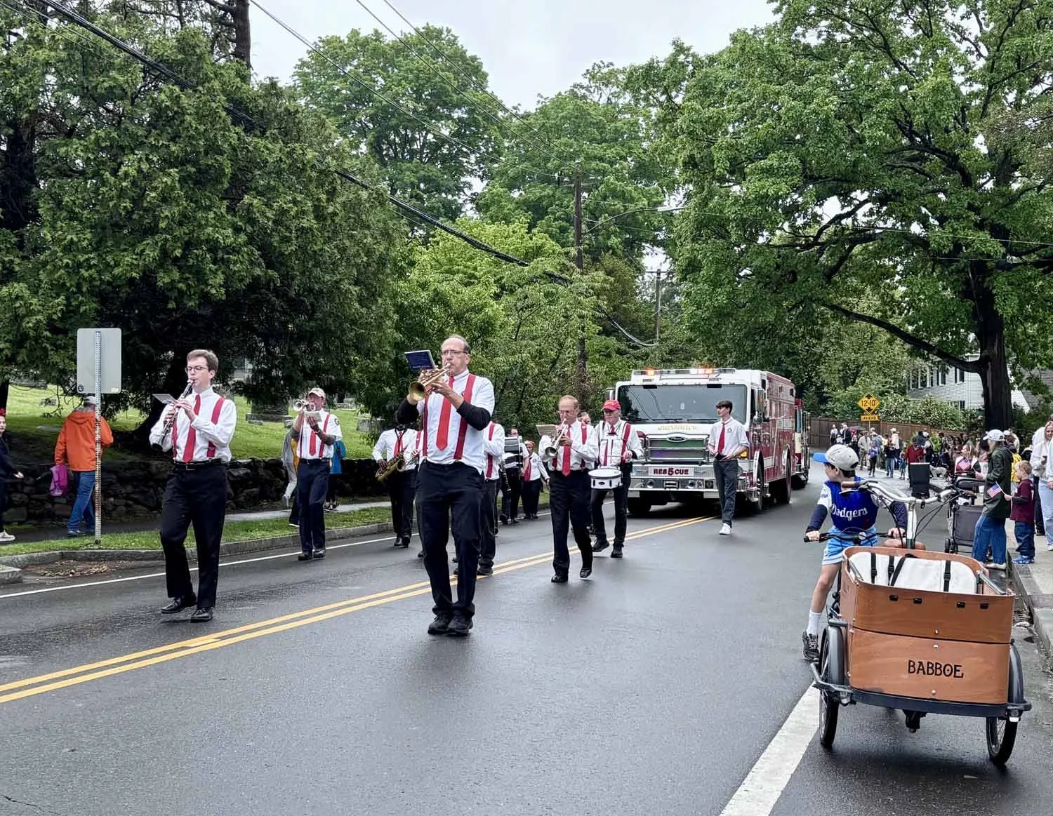 A parade featuring a marching band playing instruments like clarinets, saxophones, and drums moving down a wet street, with spectators and children on the sidewalk, some on bicycles, watching the parade on an overcast day.