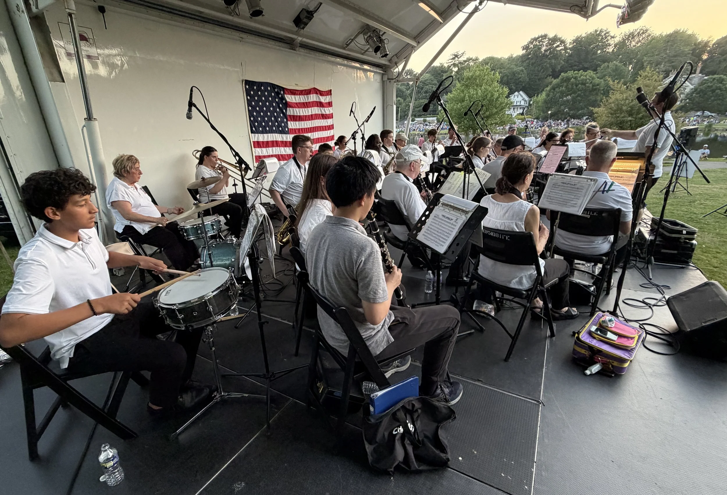 A group of musicians performing outdoors on a stage with a large American flag hanging behind them. The band includes a drummer, trumpet players, and other instrumentalists. Audience members can be seen in the background near a body of water.