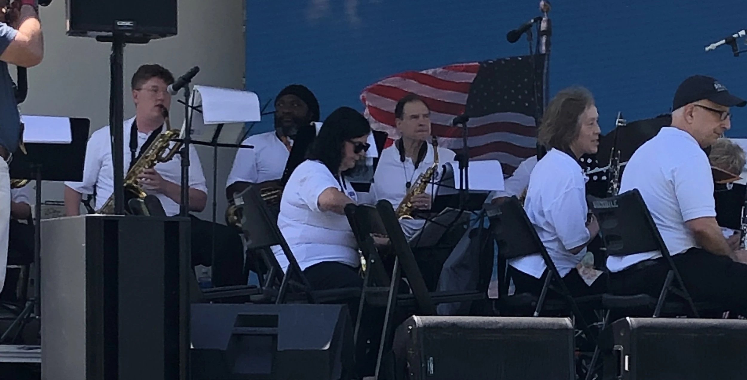 A group of musicians performing outdoors with some standing and some seated, holding instruments such as saxophones. An American flag is visible in the background, along with blue sky. Some of the musicians are wearing white shirts.