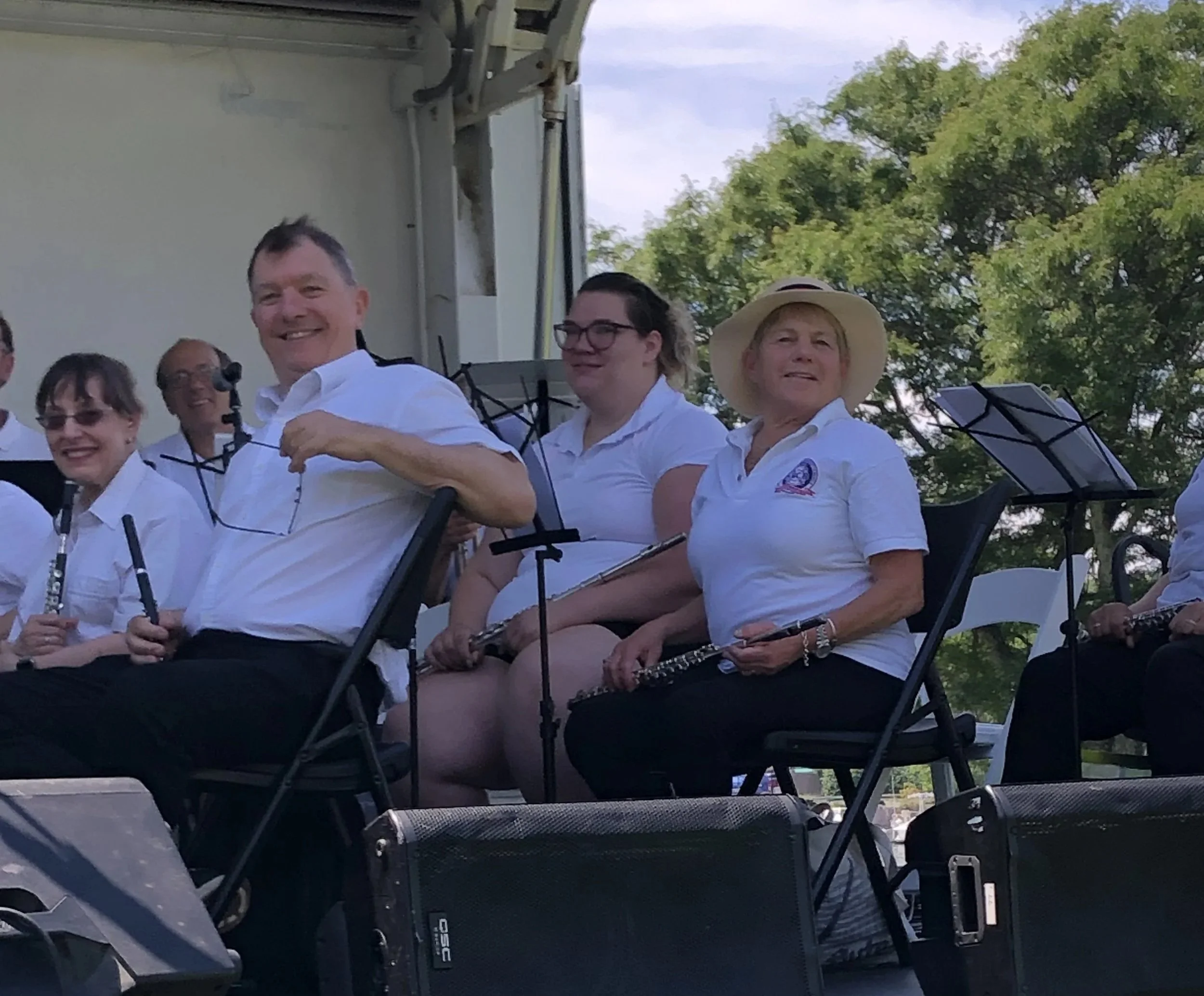 A group of people, mostly women, sitting outdoors at a concert or event, wearing white shirts, some with sunglasses and hats, with musical instruments like clarinet and music stands in front of them, green trees and a blue sky in the background.