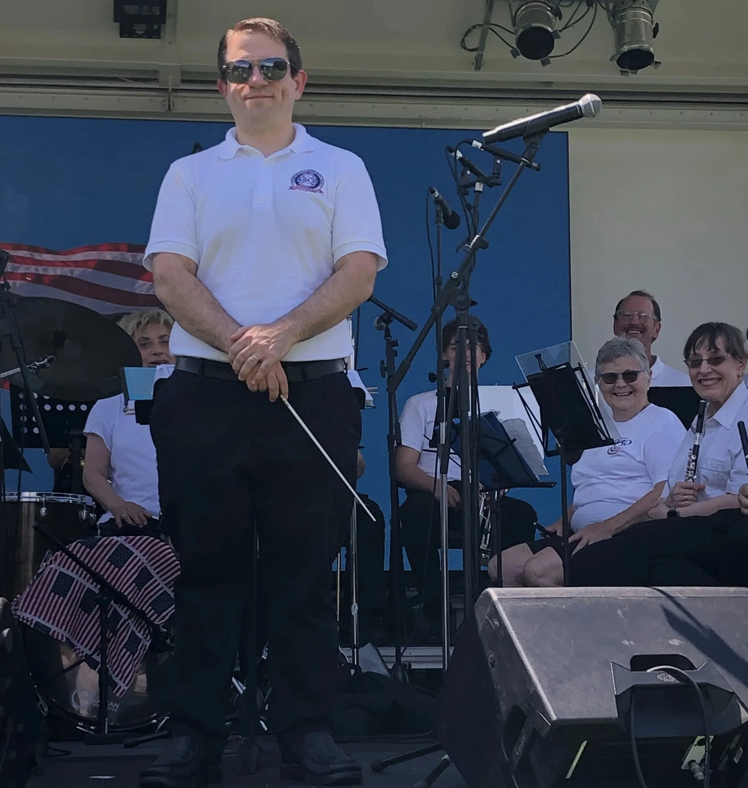 A man wearing sunglasses and a white polo shirt standing with arms crossed in front of a blue backdrop, surrounded by musicians with instruments in a performative setting.