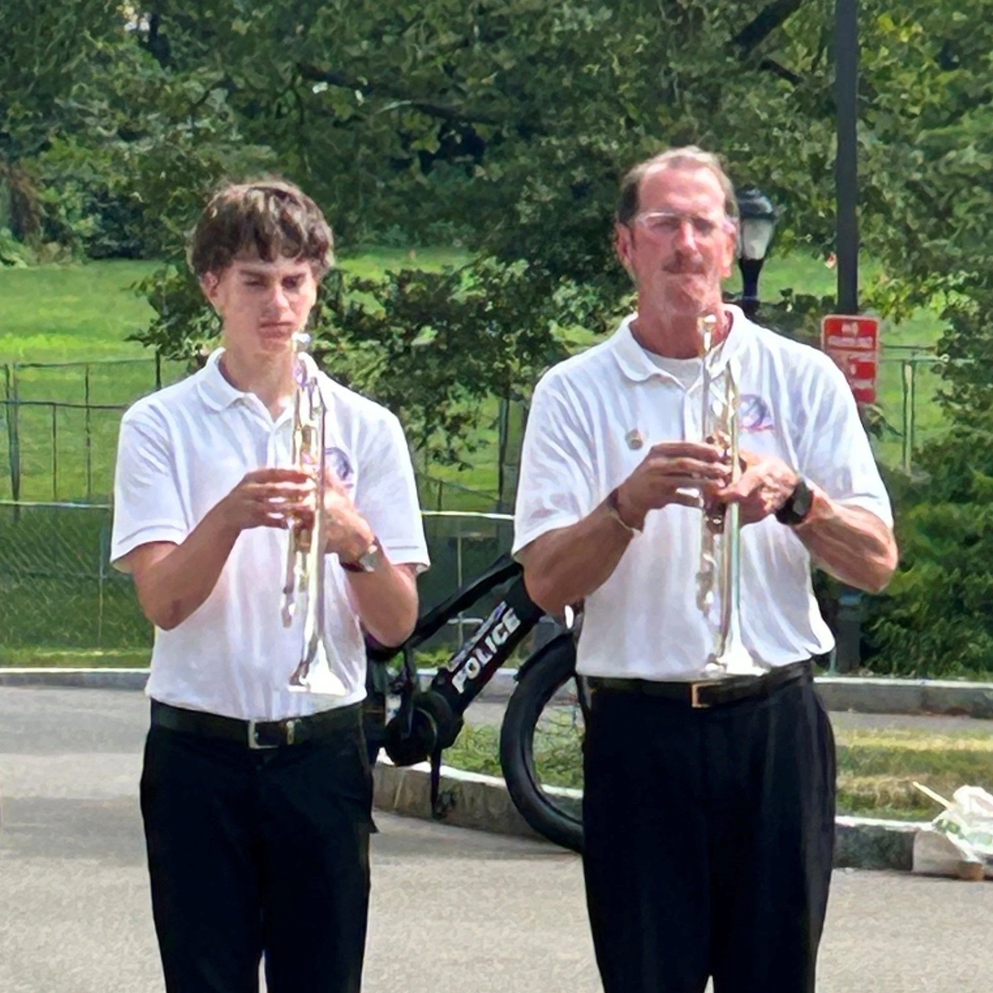 Two men standing outdoors, each holding a saxophone. They are dressed in white shirts and black pants. The background includes trees and a lamp post.