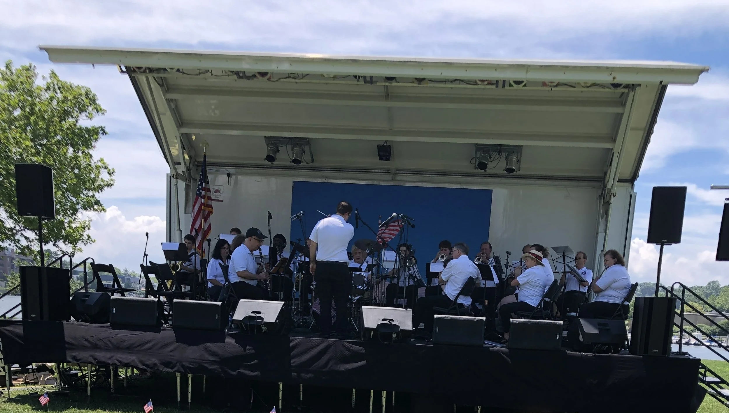 An outdoor stage with a band performing, surrounded by American flags and trees, under a partly cloudy sky.