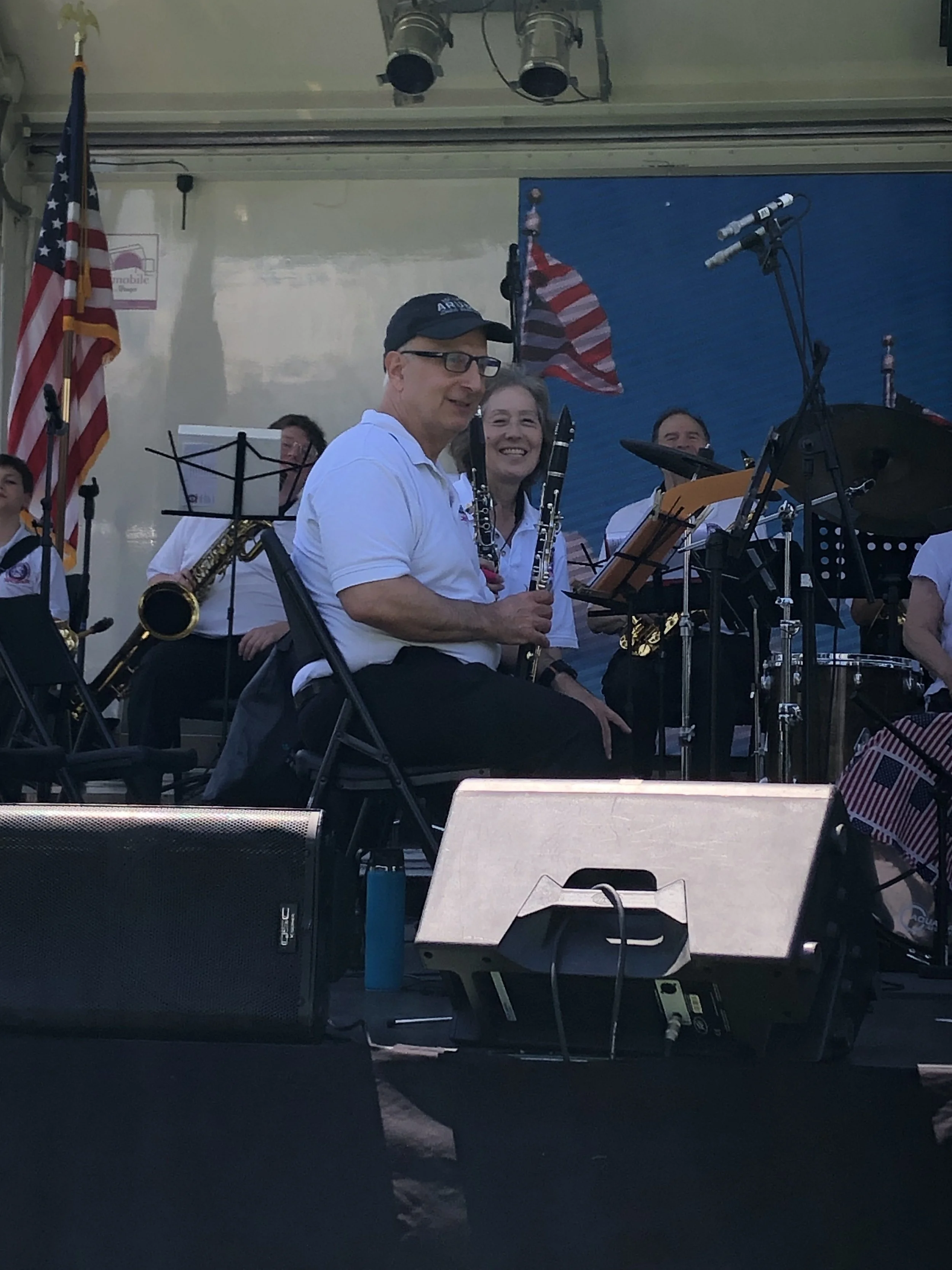 A group of musicians sitting on a stage, holding and playing wind instruments, with American flags in the background, and microphones set up in front of them.