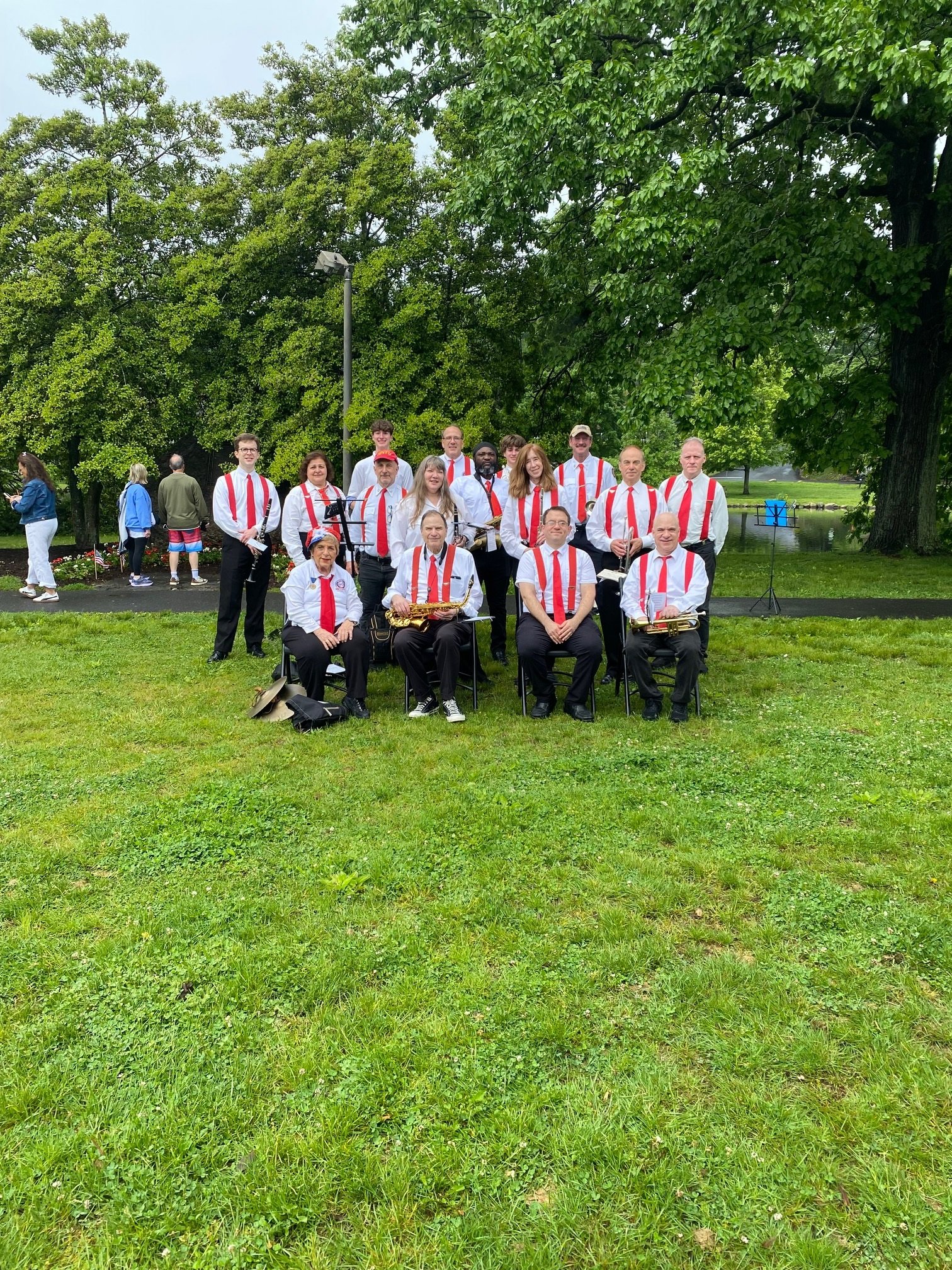 A group of people poses for a photo outdoors in a park with trees and a pond in the background. The group includes musicians dressed in white shirts with red suspenders, holding instruments, and other individuals in casual clothing.