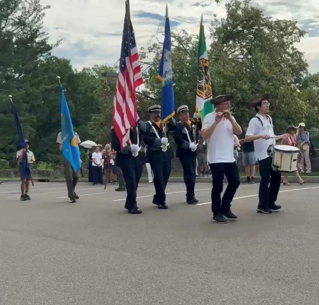 A parade with people carrying flags, including the United States flag, and musicians playing drums and wind instruments, in an outdoor setting.