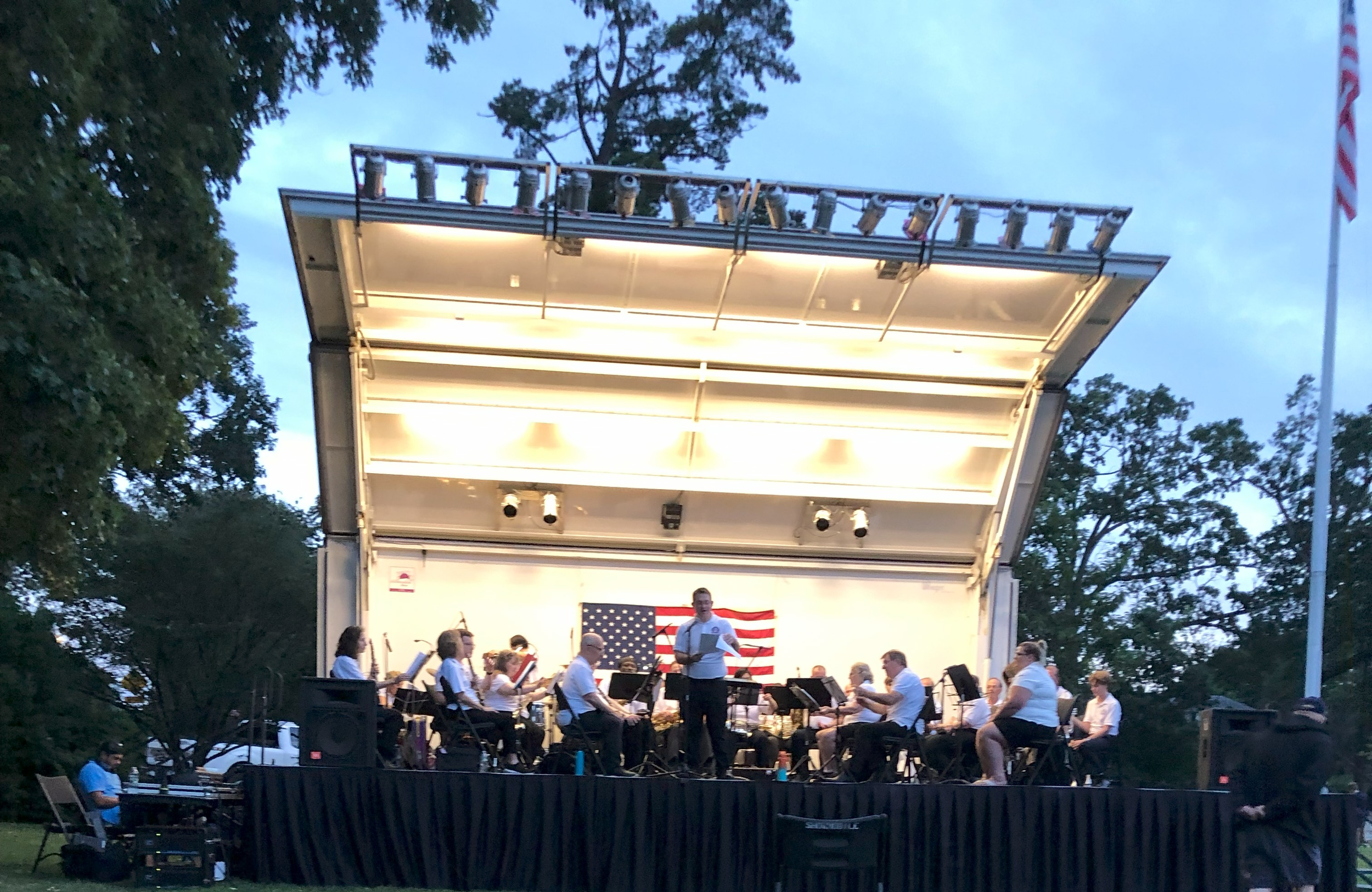 An outdoor concert stage at dusk with a band performing and a conductor leading, featuring an American flag backdrop and trees surrounding the stage.