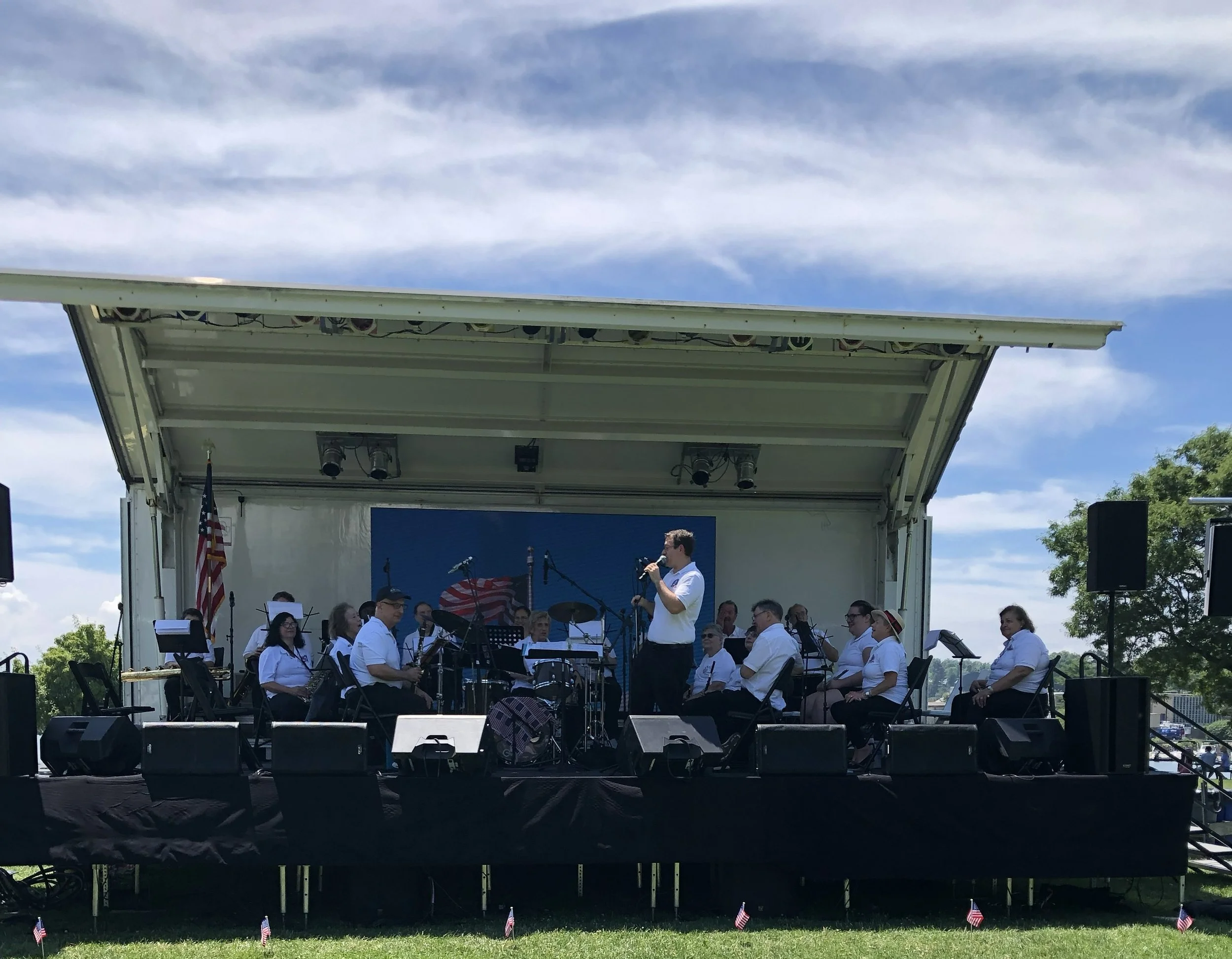 A large outdoor stage with a band performing, surrounded by small American flags on the grass, under a partly cloudy sky.