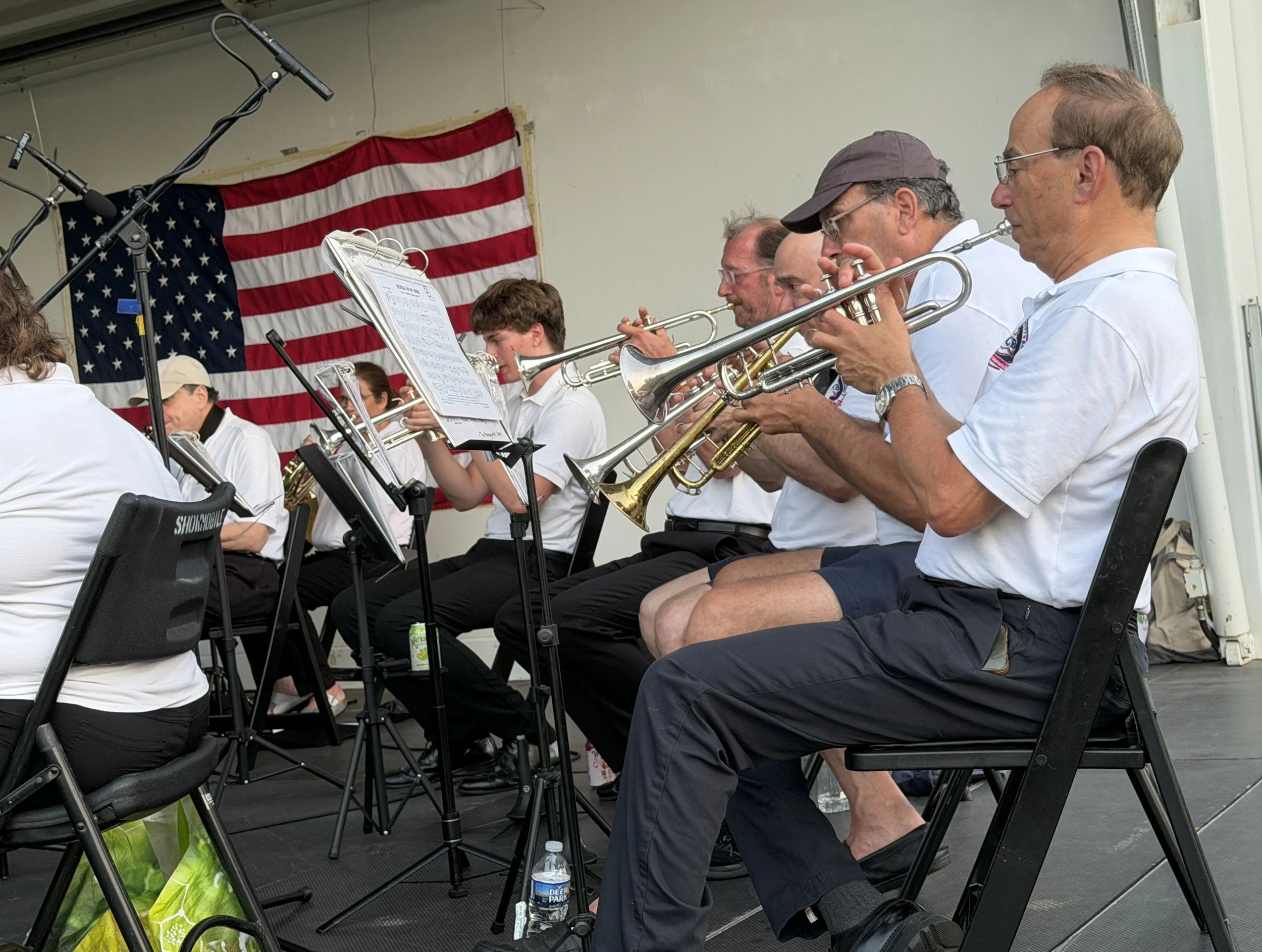 A group of musicians playing brass instruments, seated in front of an American flag, during a performance.