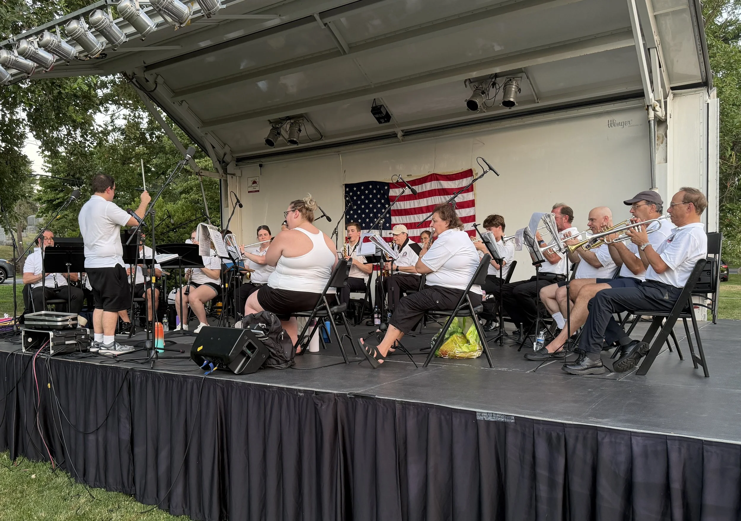 An outdoor stage with a group of musicians performing, American flag backdrop, and a conductor leading them. The stage has microphones, speakers, and stage lighting.
