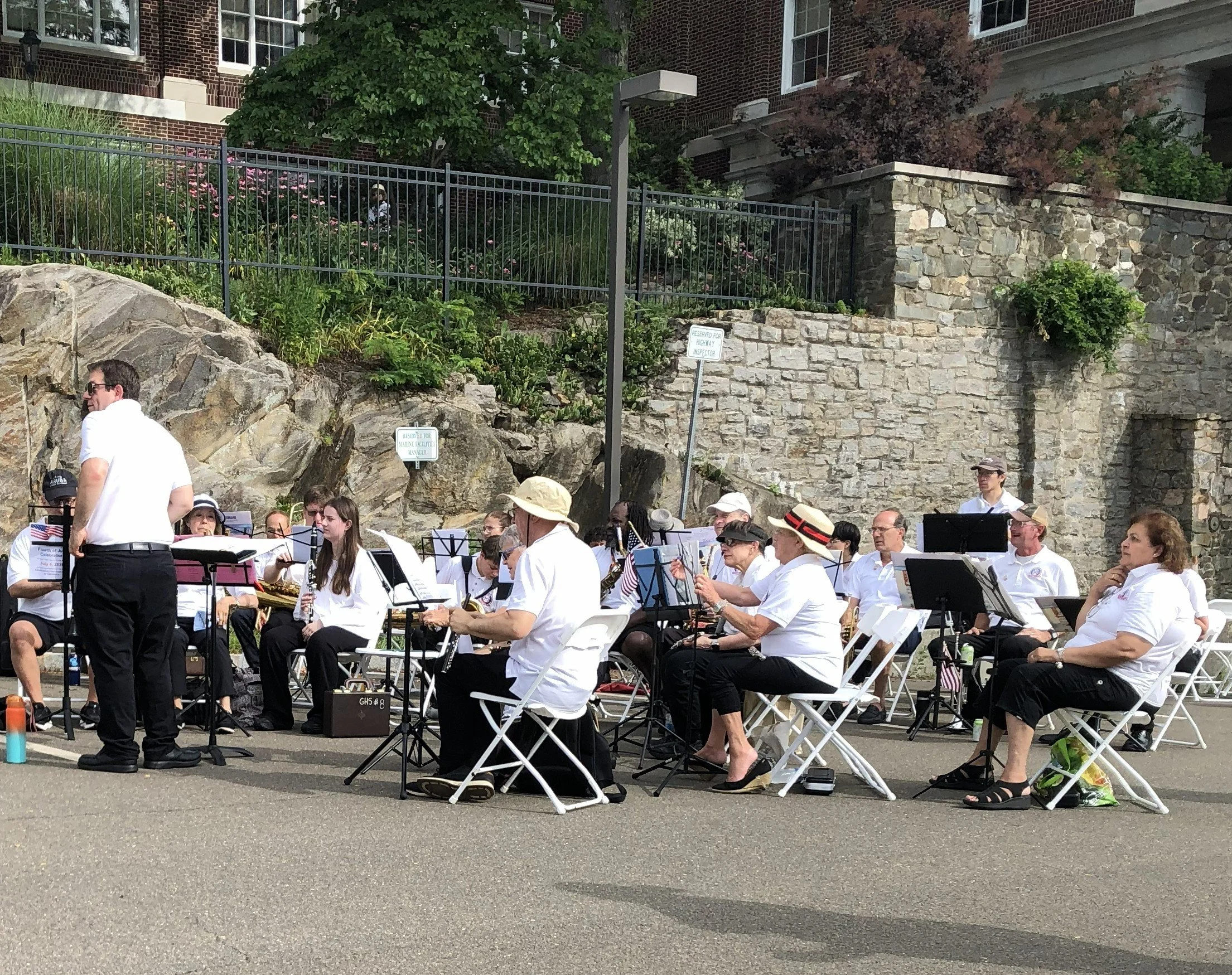 A group of musicians dressed in white shirts and black pants, sitting on chairs in an outdoor setting, performing with musical instruments such as saxophones, trumpets, and other brass instruments. They are under a clear sky, with a brick building an
