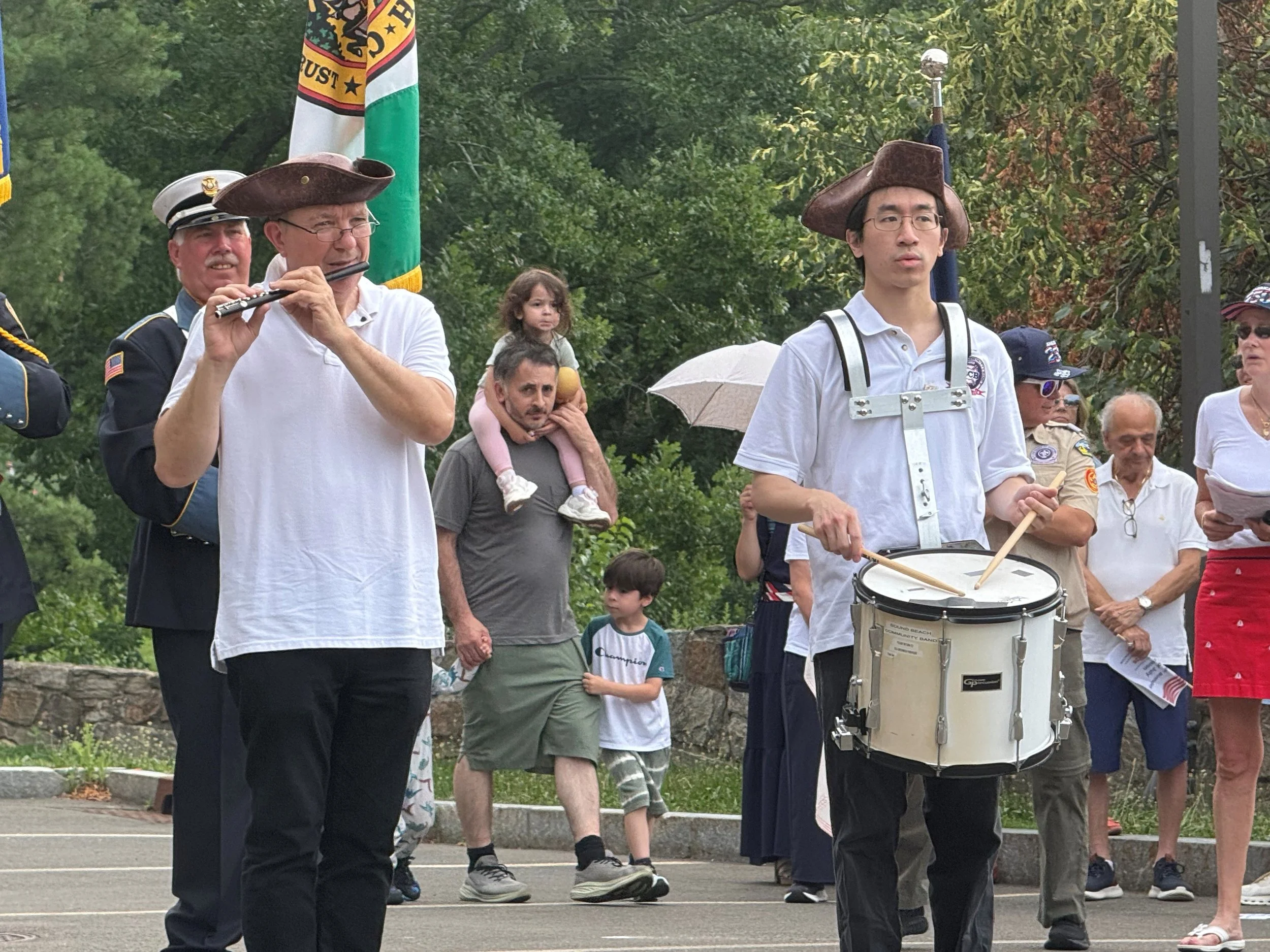 People participating in a parade or celebration, some dressed in historical or military costumes, with flags and musical instruments, in a park with green trees.