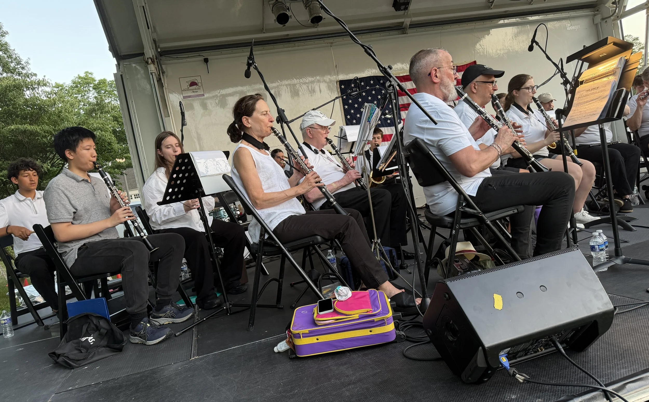 A group of musicians, including children and adults, performing on an outdoor stage with microphones and an American flag in the background during daytime.