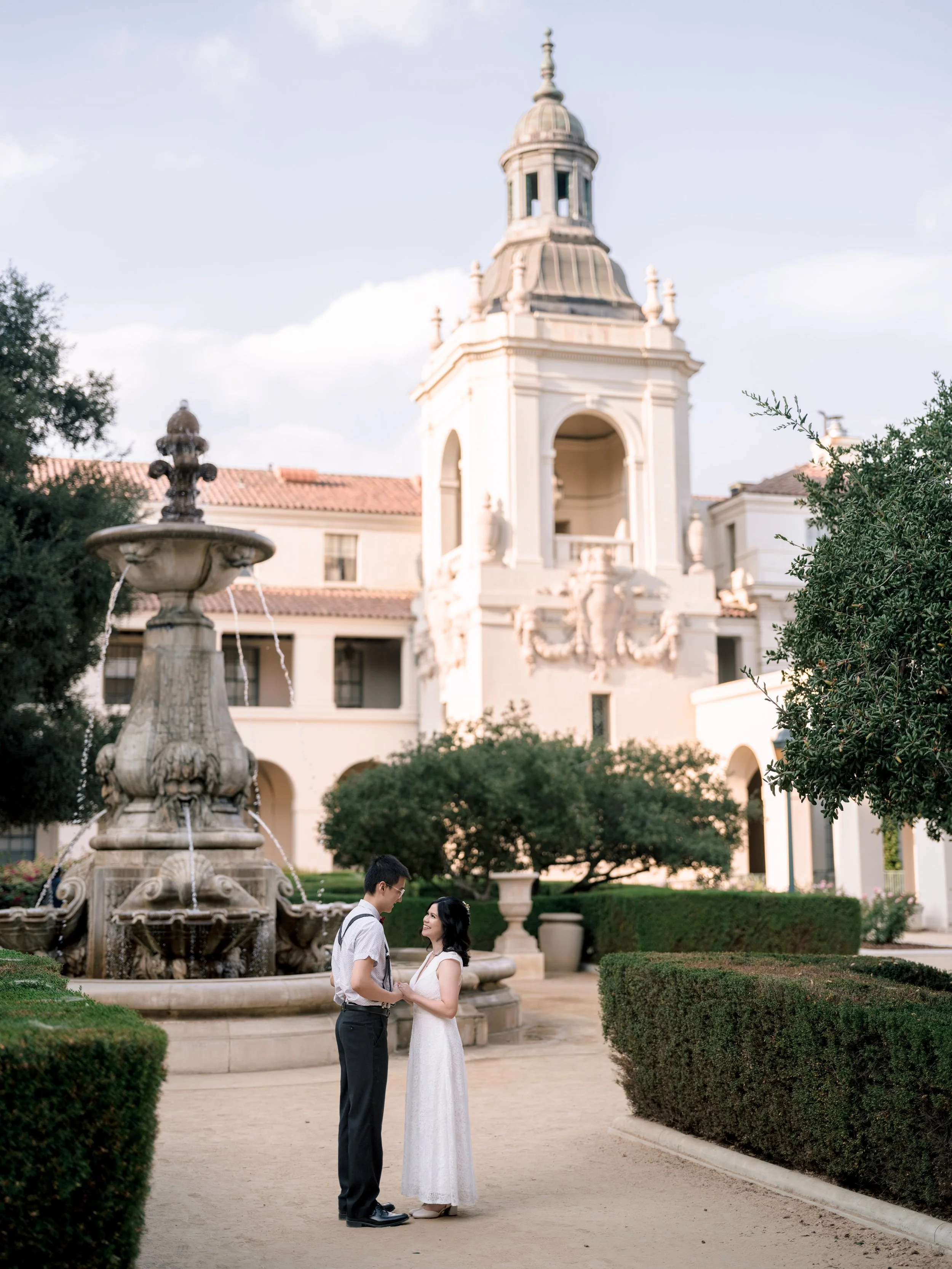 A couple holding hands faces each other in front of a fountain, with a large ornate building with a tower in the background.