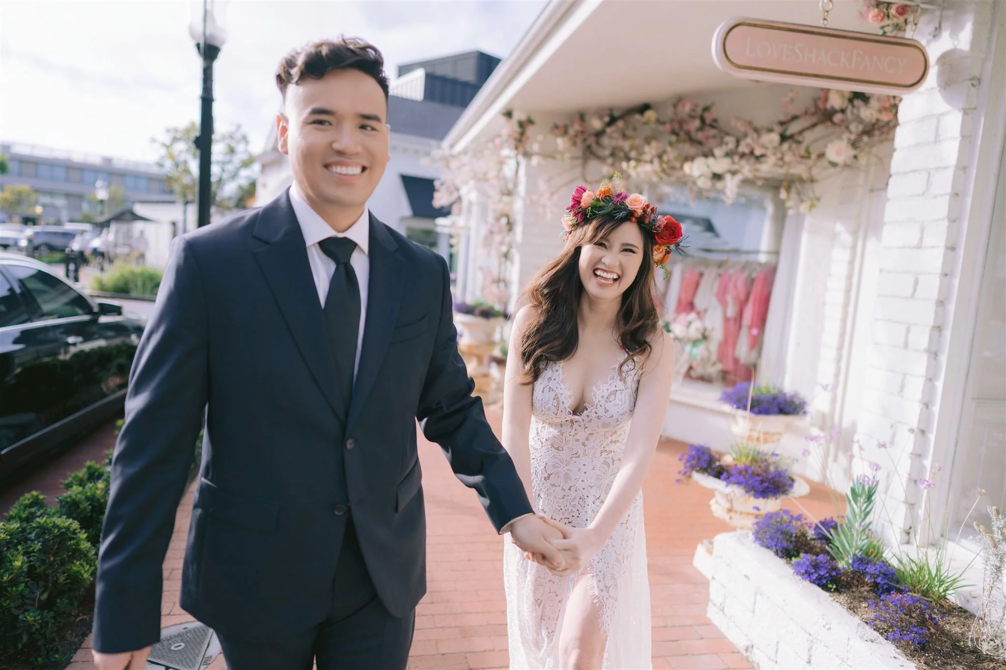 A happy couple holding hands outdoors during daytime, with the woman wearing a lace dress and floral crown, and the man dressed in a suit, in front of a shop decorated with flowers.