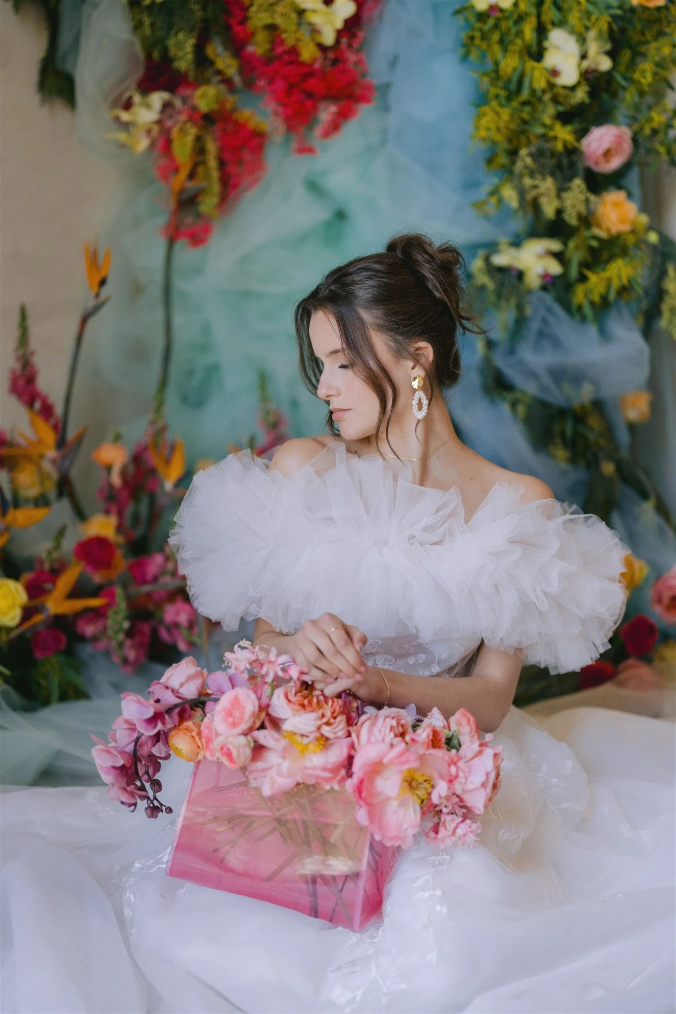 Woman in white tulle dress holding pink flowers, surrounded by colorful floral arrangements.