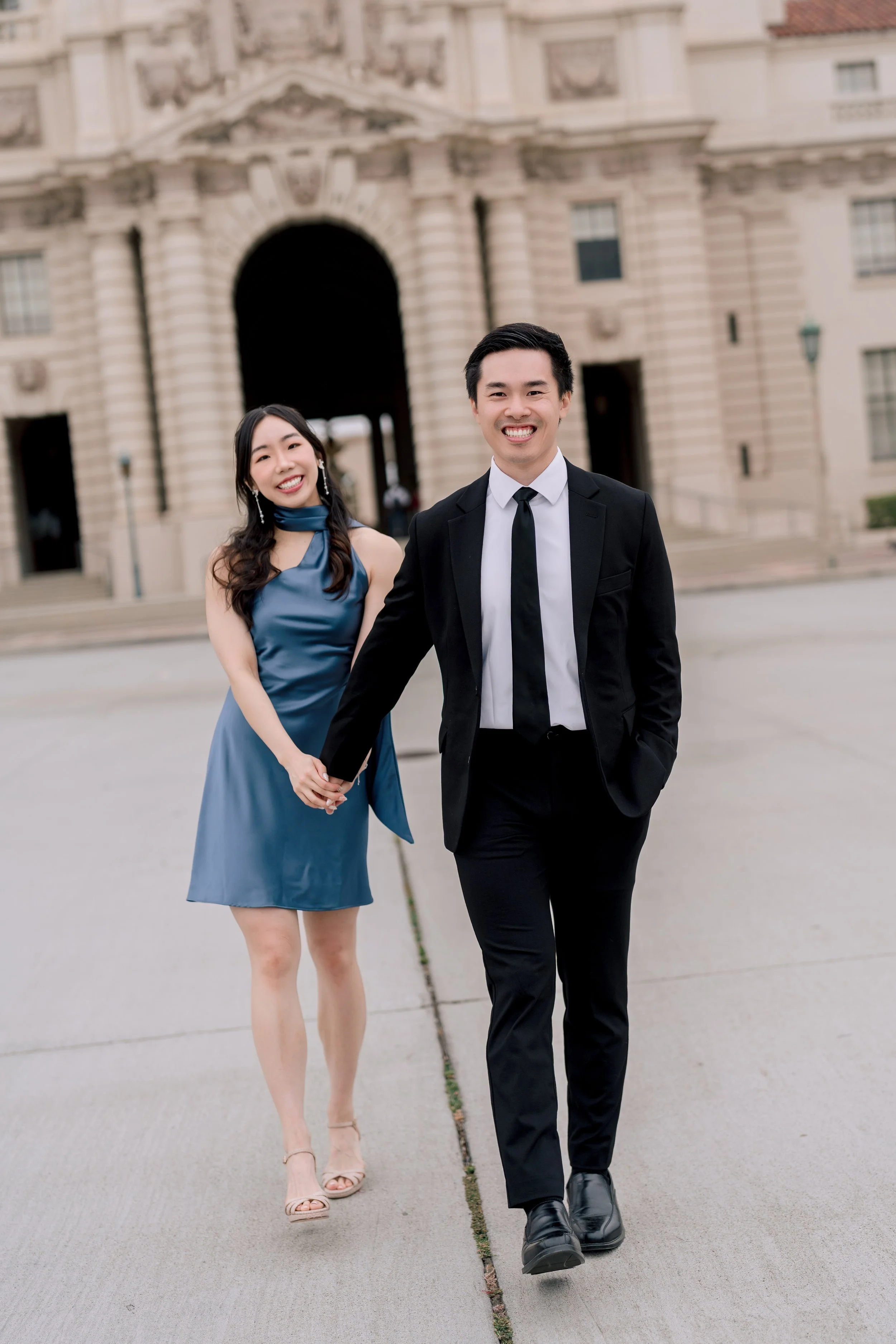A smiling young couple holding hands and walking outside in front of a large historic building. The man is dressed in a black suit with a white shirt and black tie, and the woman is wearing a blue dress with beige high heels.