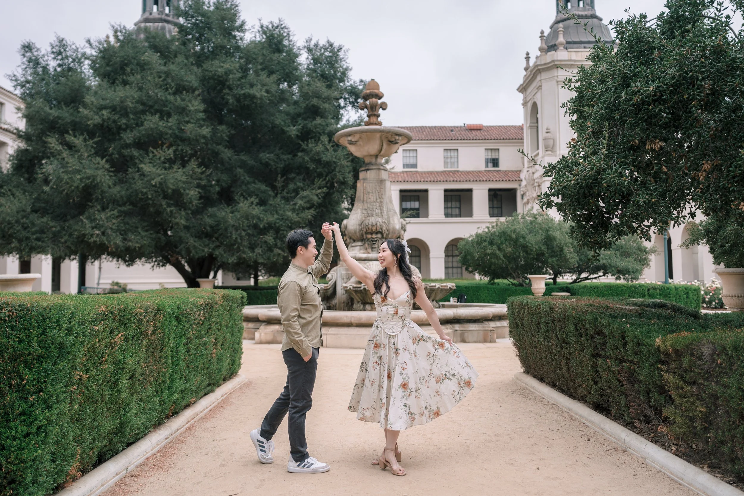 A young couple dancing and smiling in a park with a fountain, bushes, and a historic building in the background.