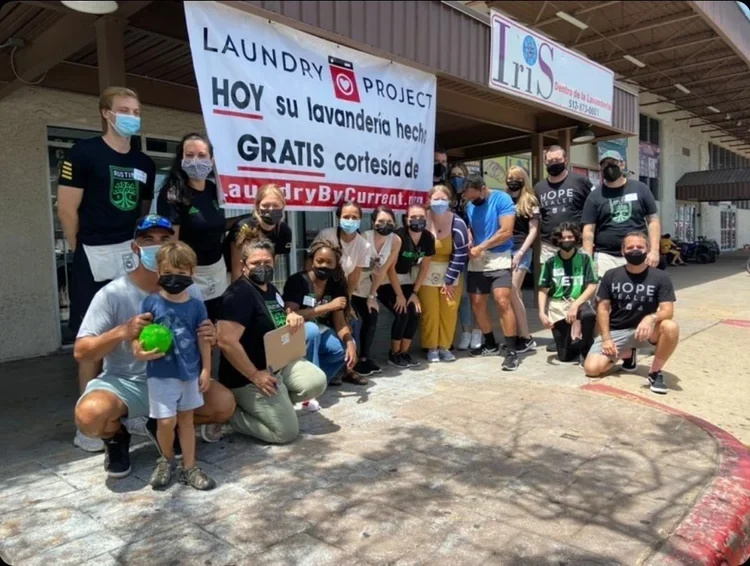 Group of people standing and kneeling outdoors near a building with a large sign about a laundry project offering free laundry services, all wearing face masks.