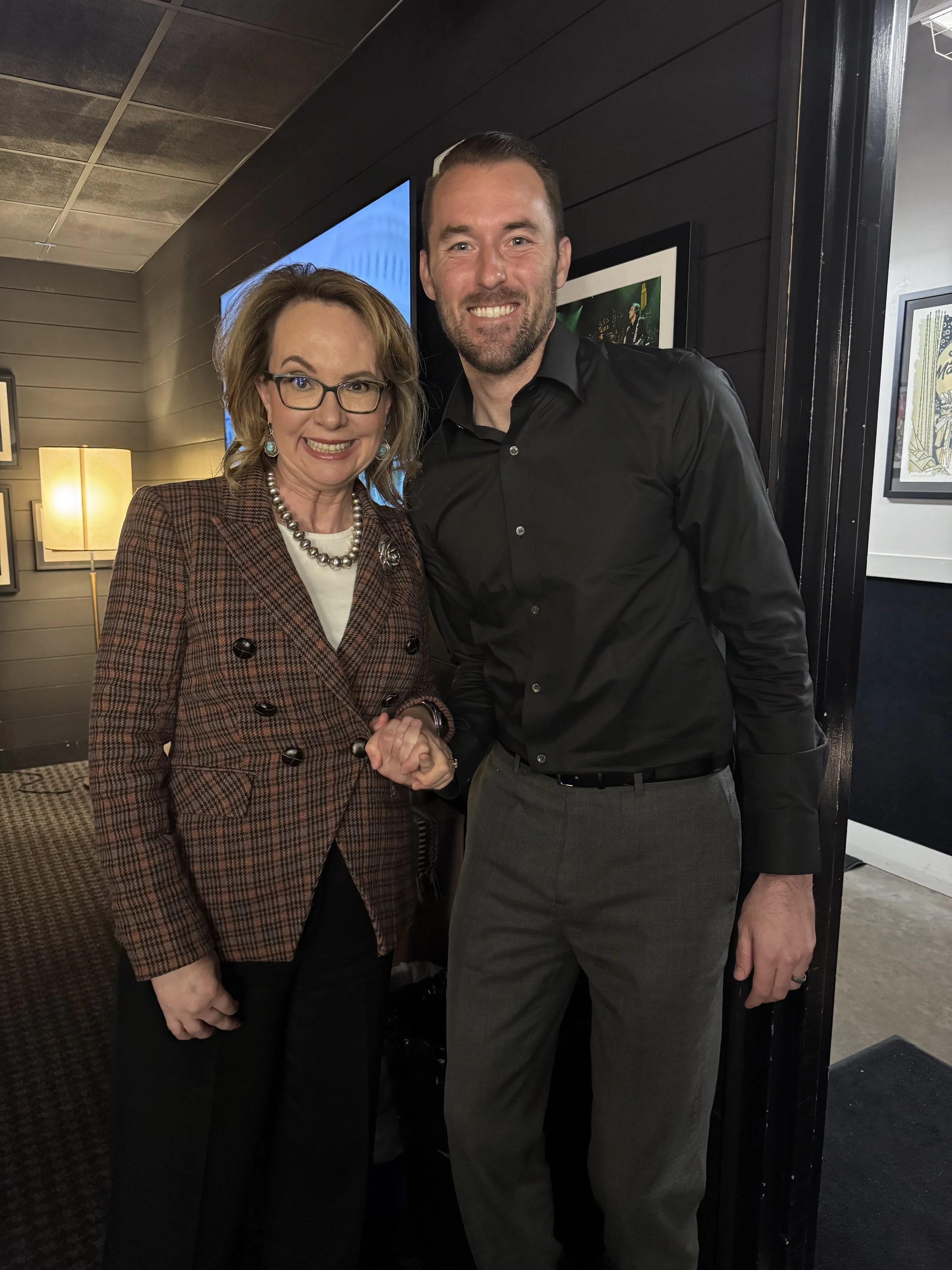 A woman and a man are posing together indoors. The woman is wearing glasses, a plaid blazer, a necklace, and is smiling. The man is dressed in a black button-up shirt and gray pants, also smiling. They are holding hands.
